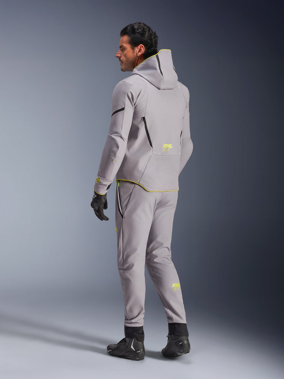 Alpinestars Aeroshell Waterproof Jacke, Mid Gray Yellow Fluo, getragen mit passender grauer Hose, Hoodie-Design mit gelben Akzenten und Logos, Rückansicht, kombiniert mit schwarzen Motorradhandschuhen und -stiefeln, moderne Motorradbekleidung