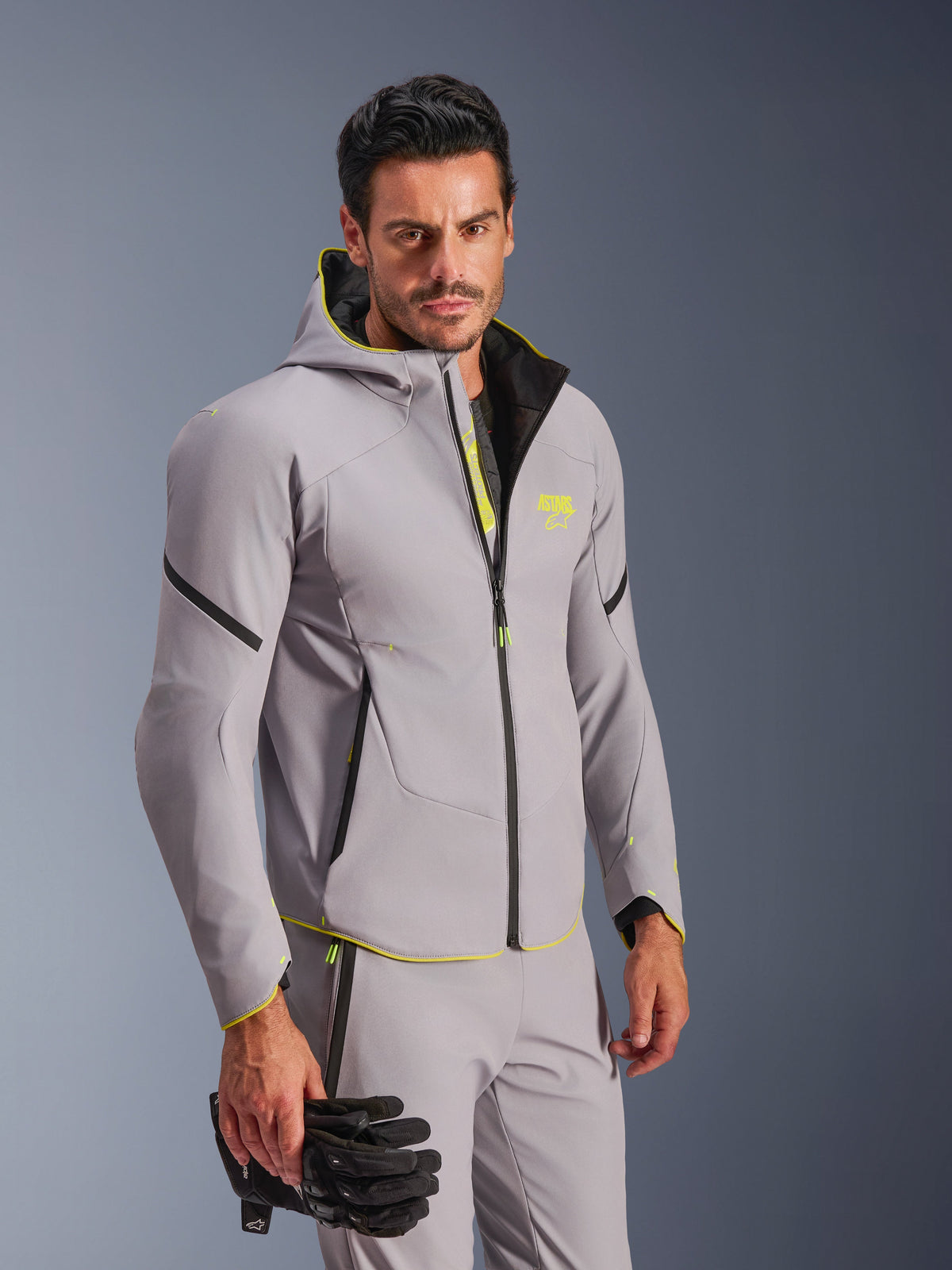 Alpinestars Aeroshell WP Jacke in Mittelgrau und Gelb Fluo, getragen von einem Mann mit passender grauer Hose und schwarzen Motorradhandschuhen, Softshell-Design mit Kapuze, neongelben Akzenten und Brust-Logo, moderne Sport-Motorradausrüstung.