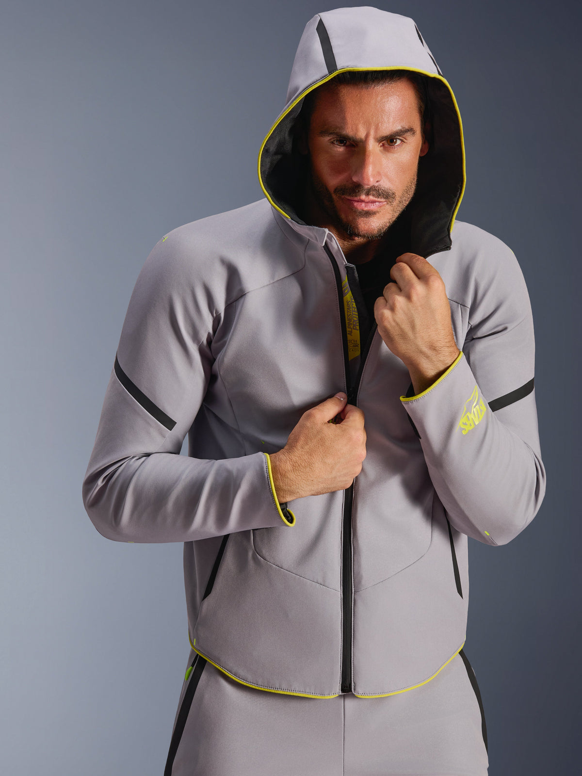 Alpinestars Aeroshell Waterproof Jacke, sportliche Softshell-Jacke mit Kapuze, Mittelgrau und Neongelb, getragen von einem männlichen Model mit aufgesetzter Kapuze, schwarze wasserdichte Reißverschlüsse und neongelbe Akzente an Ärmelbündchen und Saum, modernes Motorsport-inspiriertes Design