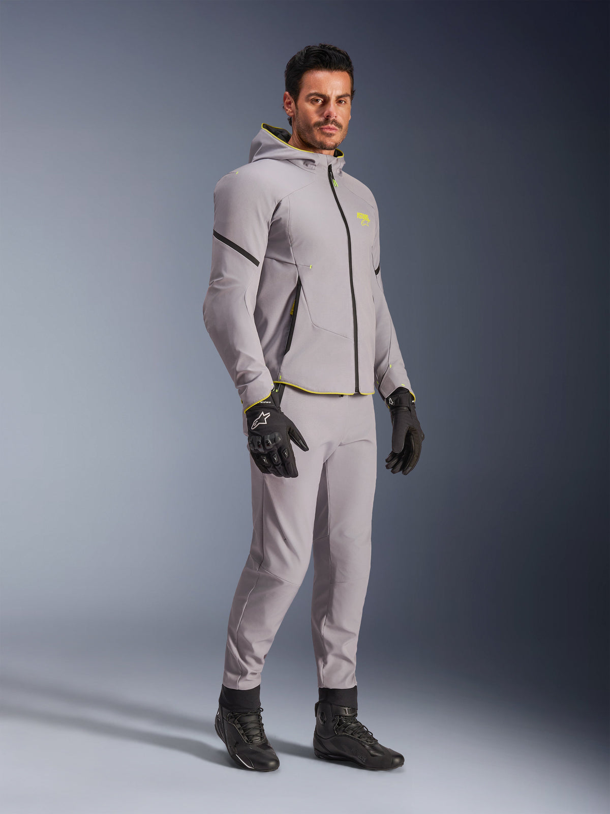 Alpinestars Aeroshell Waterproof Jacke, Mittelgrau und Yellow Fluo, getragen von einem Model mit passender grauer Hose, schwarzen Motorradhandschuhen und schwarzen Stiefeln, moderne Motorradausrüstung im Hoodie-Stil für Freizeit- und Sportfahrten.