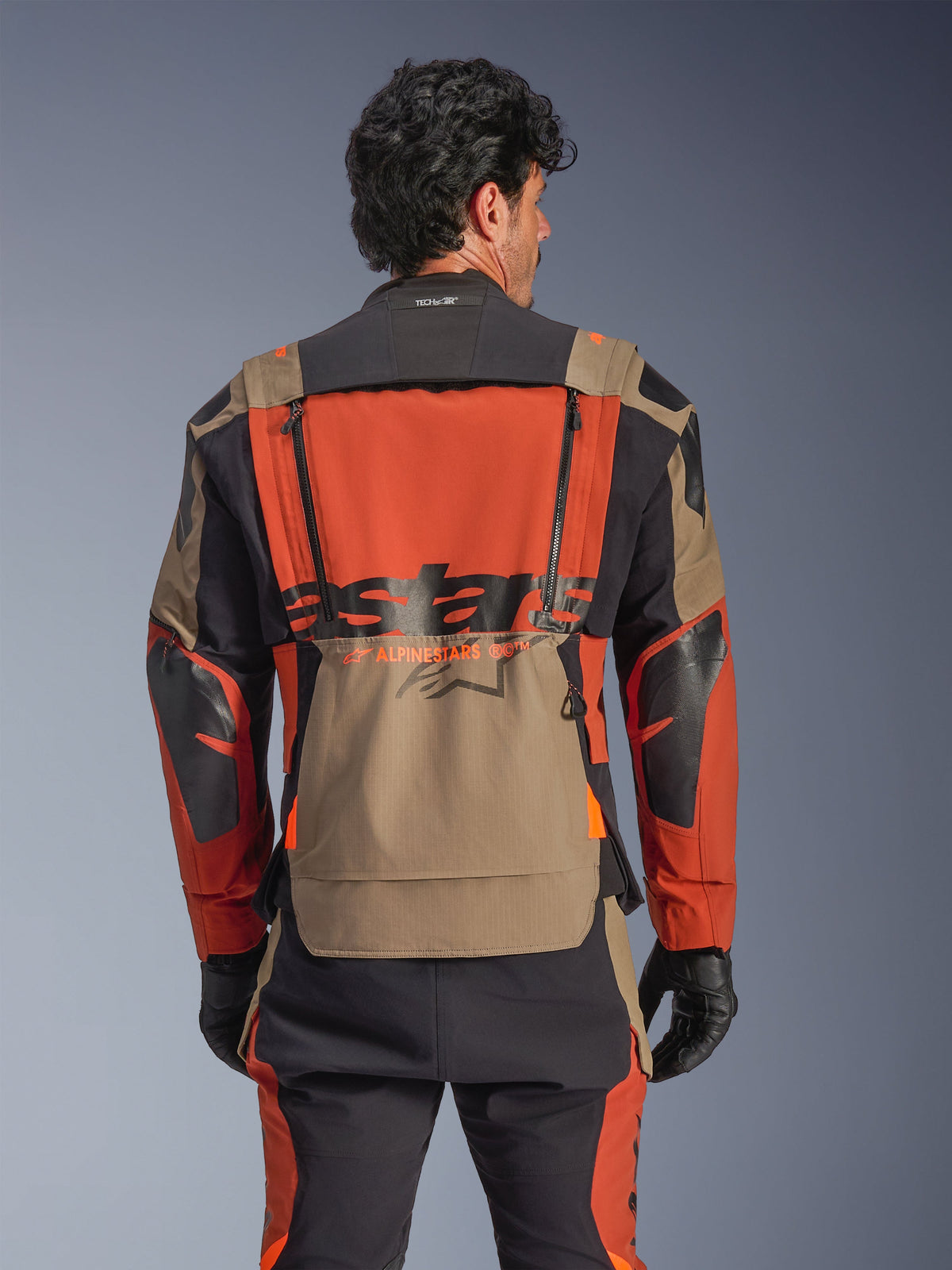 Alpinestars Halo Pro Drystar® XF Laminierte Jacke, Farbgebung Walnut Rust Black, Rückansicht, mit einem großen orangefarbenen Rückenpanel mit schwarzem ASTARS-Branding, walnussfarbener unterer Stautasche und Tech-Air® Ready Kragen, getragen mit passender Adventure-Fahrhose und schwarzen Handschuhen