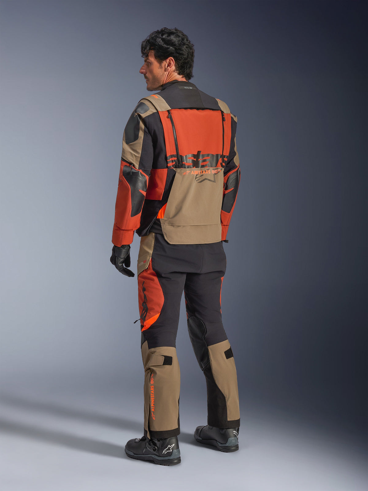 Alpinestars Halo Pro Drystar® XF Laminierte Jacke, Adventure-Motorradjacke in Walnut Rust Black, Rückansicht getragen mit passender Hose und schwarzen Stiefeln, mit technischem 3L-Stretchmaterial, modularen Belüftungspaneelen und orangefarbenem Branding auf der Rückseite
