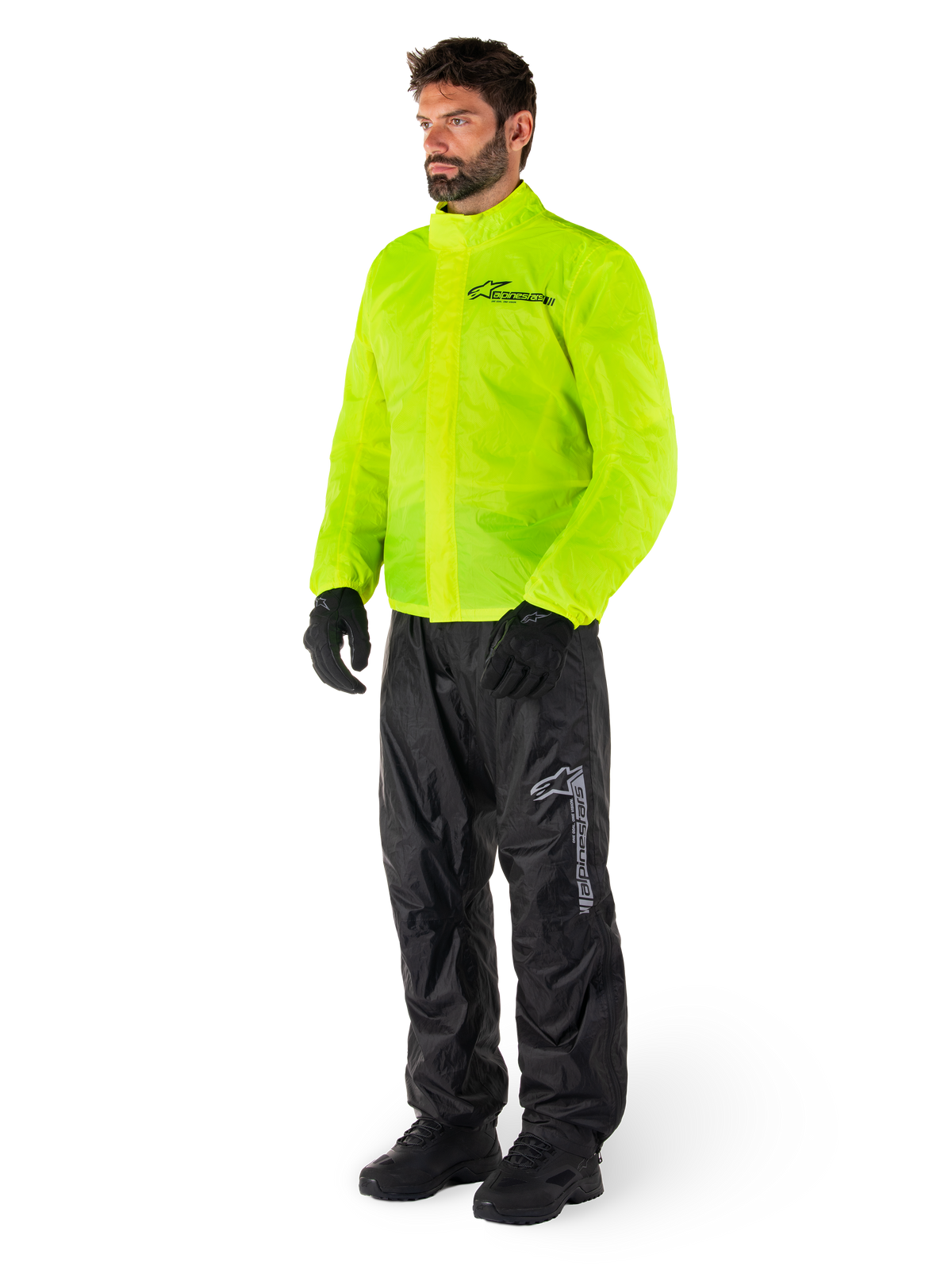 Mann trägt Alpinestars Hurricane Rain V2 Jacke in Neongelb und Hurricane Rain V2 Hose in Schwarz, hochsichtbare wasserdichte Motorrad-Regenbekleidung, mit reflektierenden Logos auf der Brust und am Oberschenkel, komplettes Outfit mit schwarzen Handschuhen und Stiefeln.