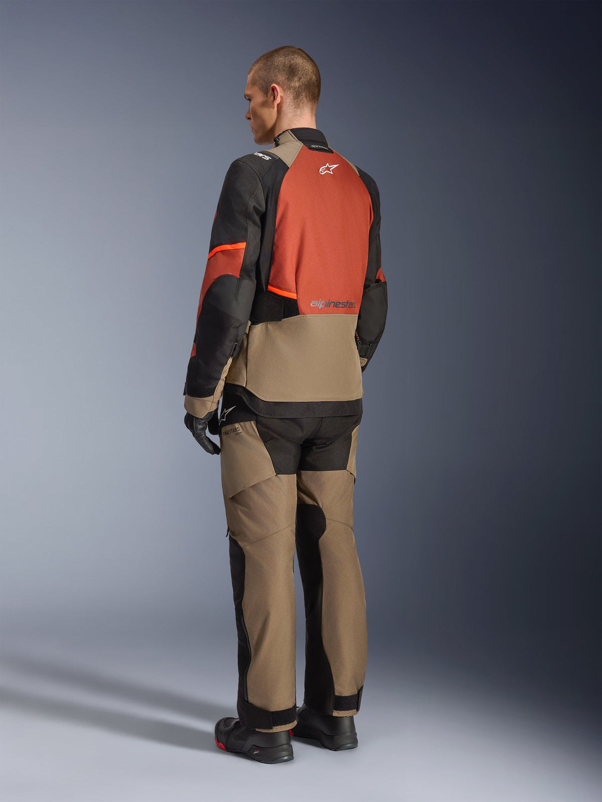 Alpinestars Andes V4 Drystar® Jacke in Walnut Rust Black, Modell in der Rückansicht mit passender Textilhose, schwarzen Handschuhen und Stiefeln, technisches Multi-Panel-Design für Adventure-Touring und Alpinestars-Branding.