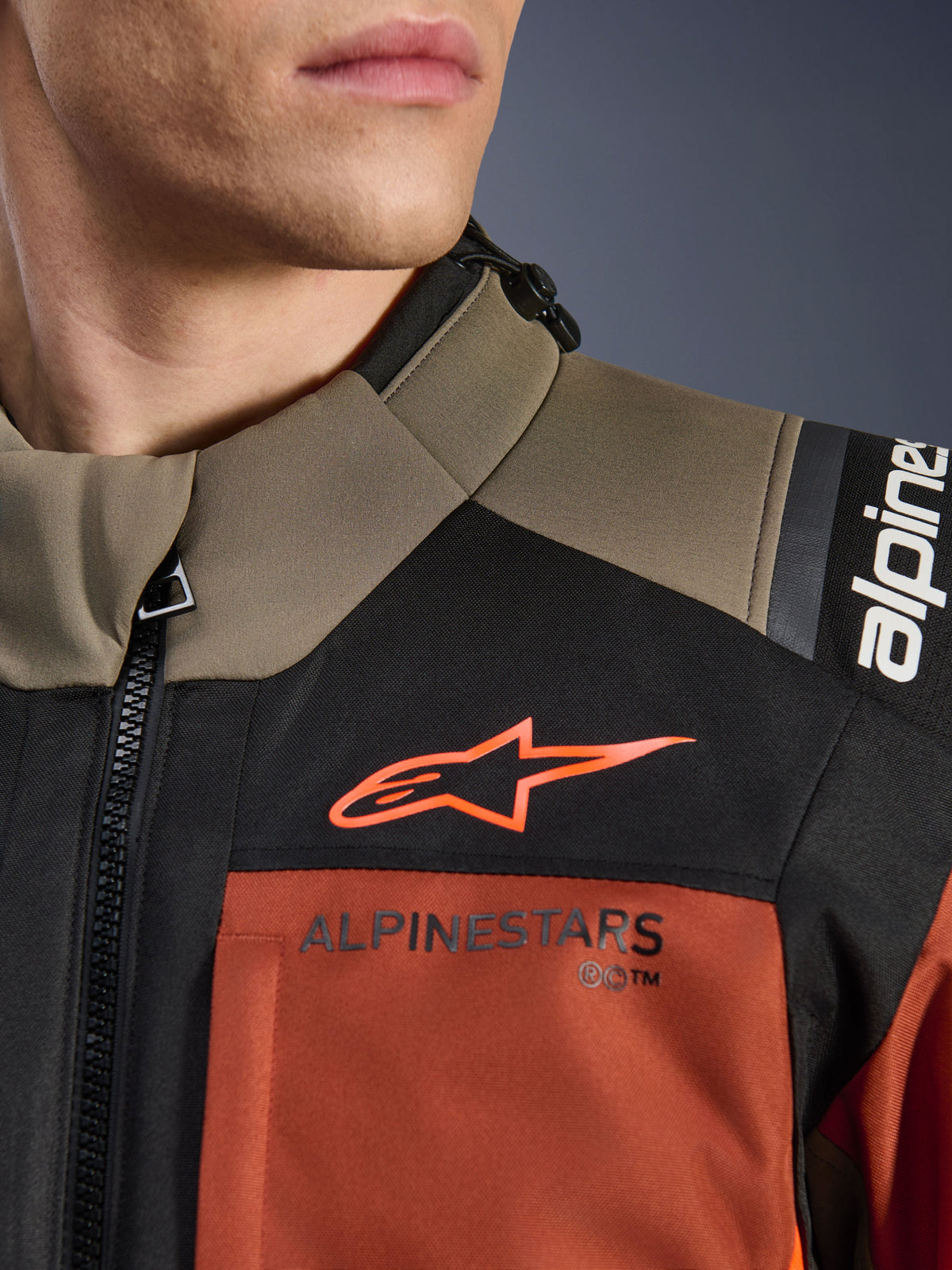 Alpinestars Andes V4 Drystar® Jacke in Walnut Rust Black, Nahaufnahme des oberen Brustbereichs und der Schulter, orangefarbenes A-Star-Logo auf schwarzem Panel und Alpinestars-Schriftzug auf rostfarbenem Abschnitt, Detail einer Adventure-Motorradausrüstung