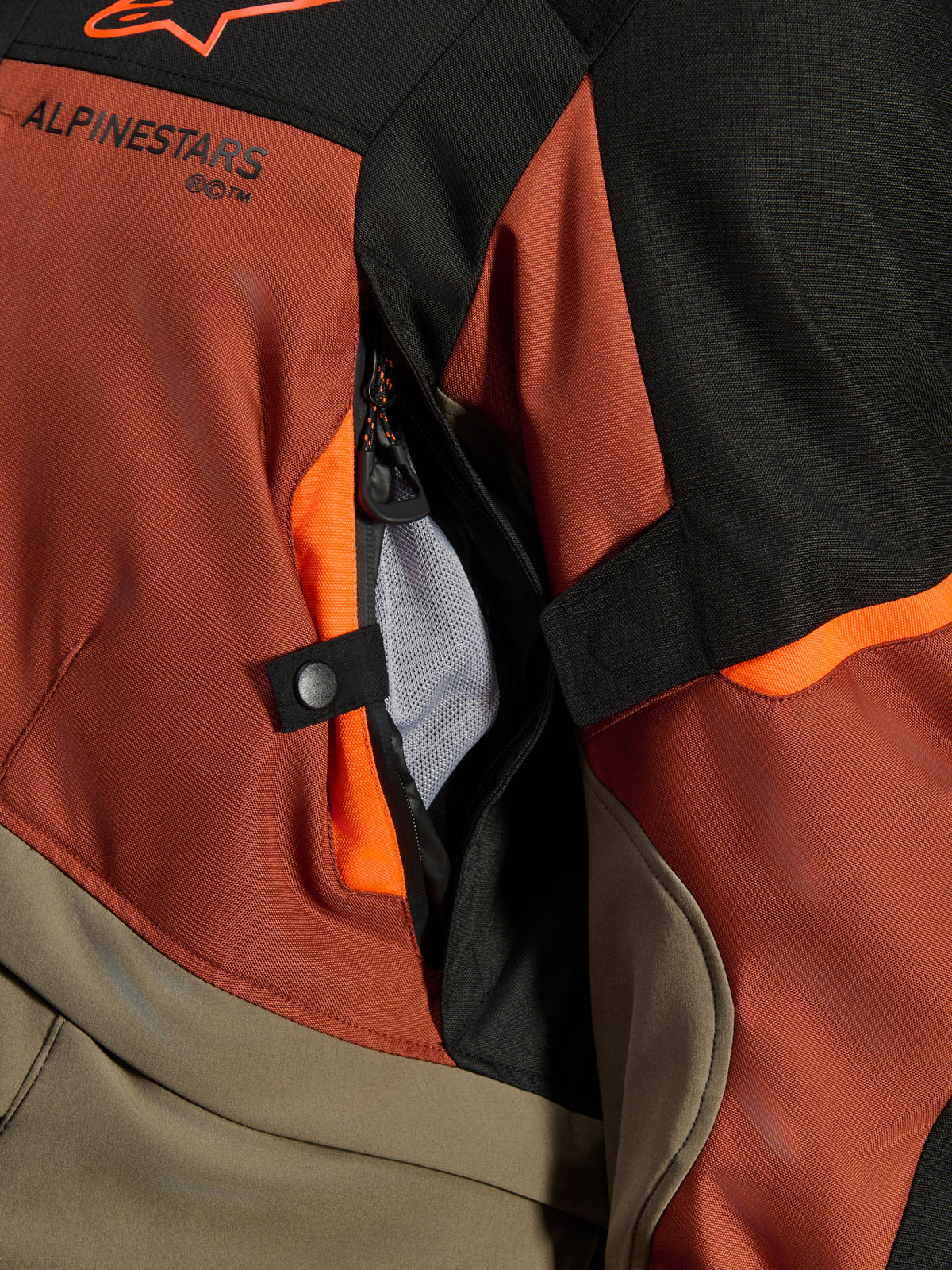 Alpinestars Andes V4 Drystar® Jacke, Walnut Rust Black, Nahaufnahme der direkten Belüftungsöffnung mit Mesh-Futter, strapazierfähiges Textilmaterial mit orangefarbenen Akzenten, Druckknopf und Alpinestars Logo-Detail