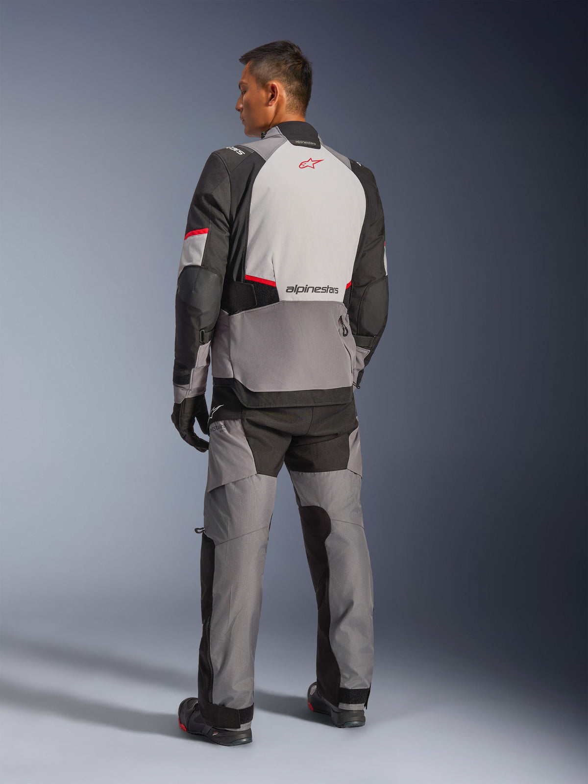 Veste Alpinestars Andes V4 Drystar®, gris foncé et gris glacier avec accents noirs et rouge vif, vue arrière montrant un panneau gris clair avec logo étoile rouge, associée à un pantalon d'aventure assorti et des gants noirs, équipement technique de moto toutes saisons