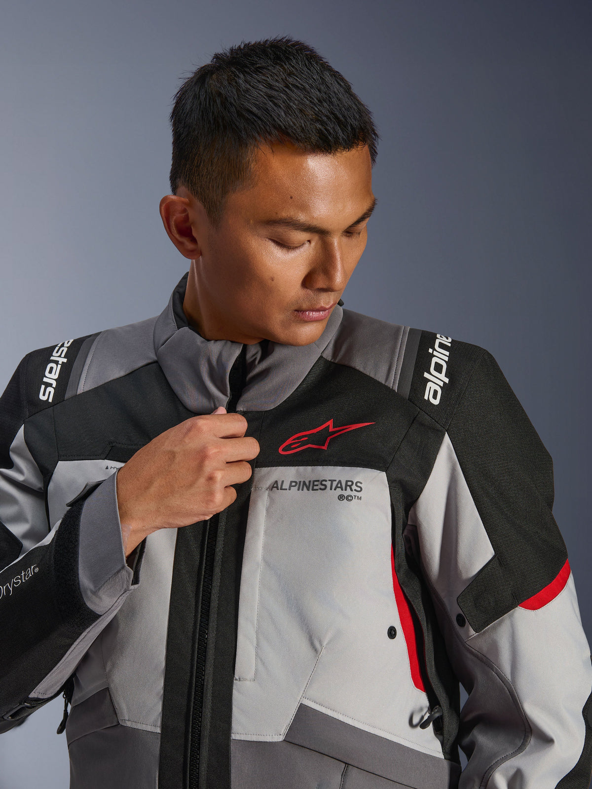 Mannequin portant la veste Alpinestars Andes V4 Drystar®, gris foncé et gris glace avec des accents noirs et rouge vif, ajustant la fermeture éclair frontale, arborant le logo Astars rouge et le marquage Drystar®, style touring aventure