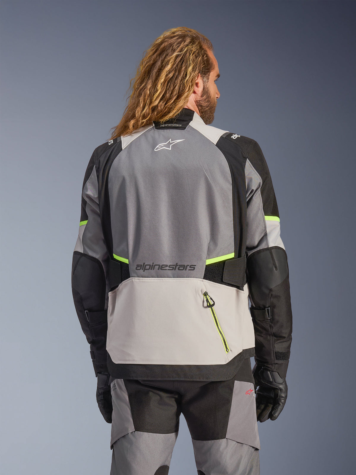 Veste Alpinestars Andes V4 Drystar®, vue arrière d'une veste de moto d'aventure en gris foncé, gris glacier et noir avec des accents jaune fluo, dotée de panneaux textiles renforcés et du marquage Alpinestars, portée avec un pantalon de moto assorti et des gants noirs