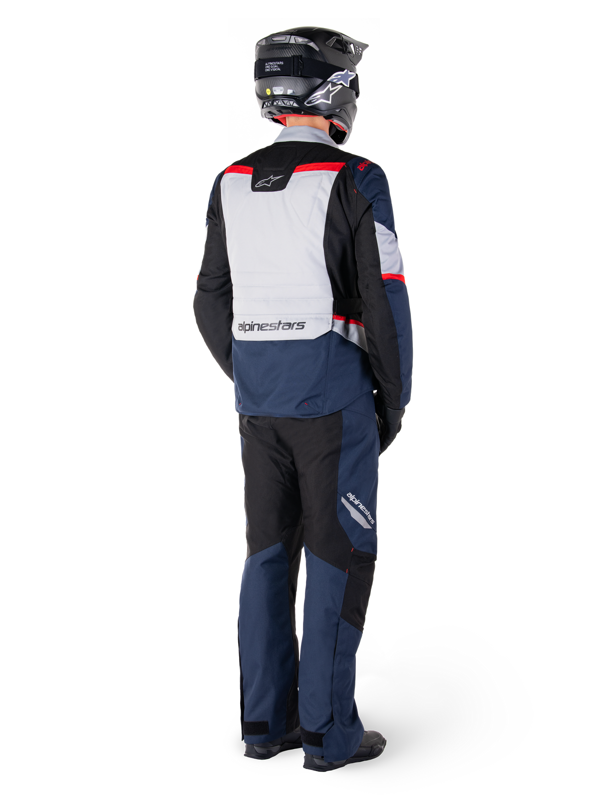 Vue arrière d'un pilote portant la veste imperméable Alpinestars ST-1 WP en bleu foncé, noir et rouge vif, associée à un pantalon imperméable assorti et un casque noir, présentant un panneau dorsal gris proéminent avec le logo Alpinestars et un design de tourisme d'aventure.