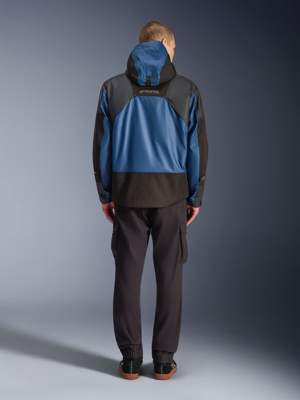 Alpinestars Mospher Waterproof jas in zwart en Dark Petrol, achteraanzicht op model, gecombineerd met een zwarte cargobroek, voorzien van tactische ripstop-constructie en MO.ST.EQ.-branding op de rug