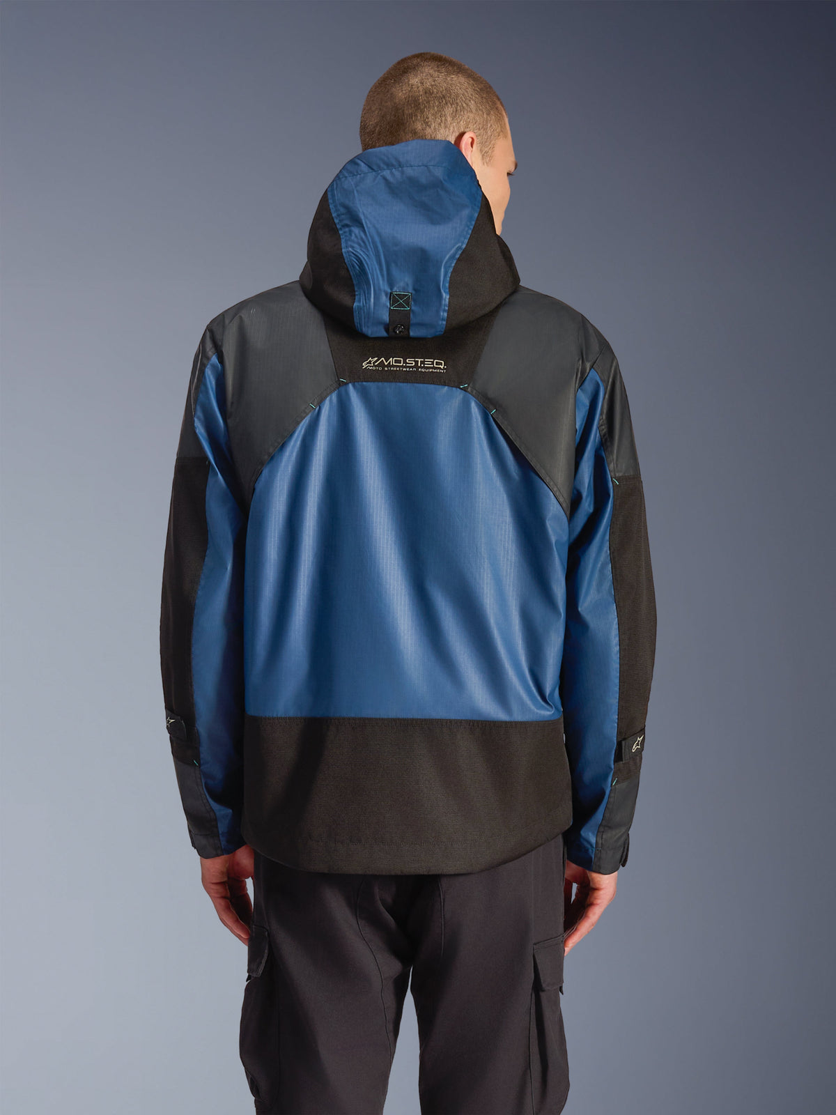 Alpinestars Mospher Waterproof Jas in Black Dark Petrol, achteraanzicht van een motorparka met blauwe ripstop panelen en zwarte verstevigde delen, MO.ST.EQ. branding op de bovenrug, modern urban tactisch ontwerp