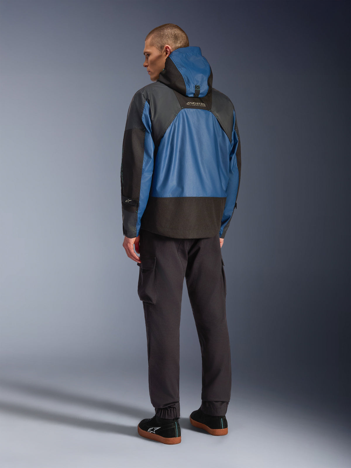 Achteraanzicht van een mannelijk model die de Alpinestars Mospher Waterproof Jacket in Black en Dark Petrol draagt, voorzien van een ontwerp met capuchon en tactische ripstop-constructie, gecombineerd met een zwarte cargobroek en sneakers.