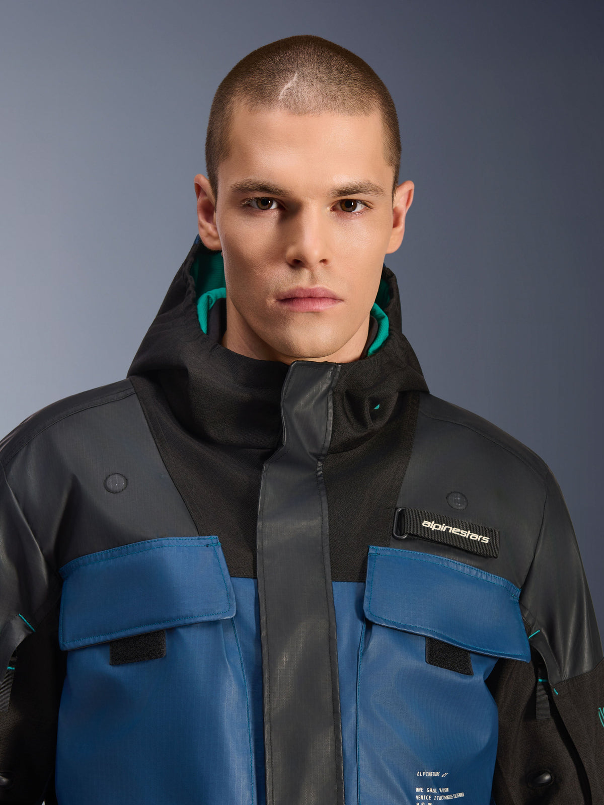 Alpinestars Mospher Waterproof Jas, Zwart en Dark Petrol, close-up van een mannelijk model dat de op utility geïnspireerde parka draagt, voorzien van donkerblauwe borstzakken, een zwarte capuchon met teal voering en Alpinestars-branding op de borst.