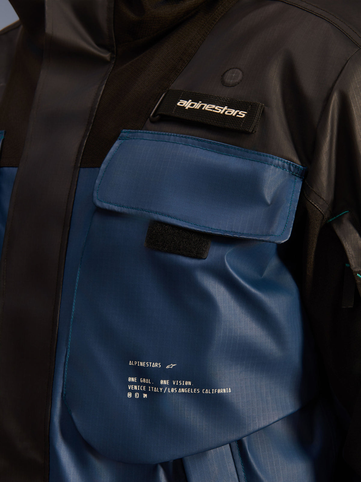 Alpinestars Mospher Waterproof jas, detailopname van de borstzak, Black en Dark Petrol, duurzame ripstop-stof met bedrukte 'One Goal. One Vision.' branding, functioneel ontwerp met praktische zakken en Alpinestars-logolabel