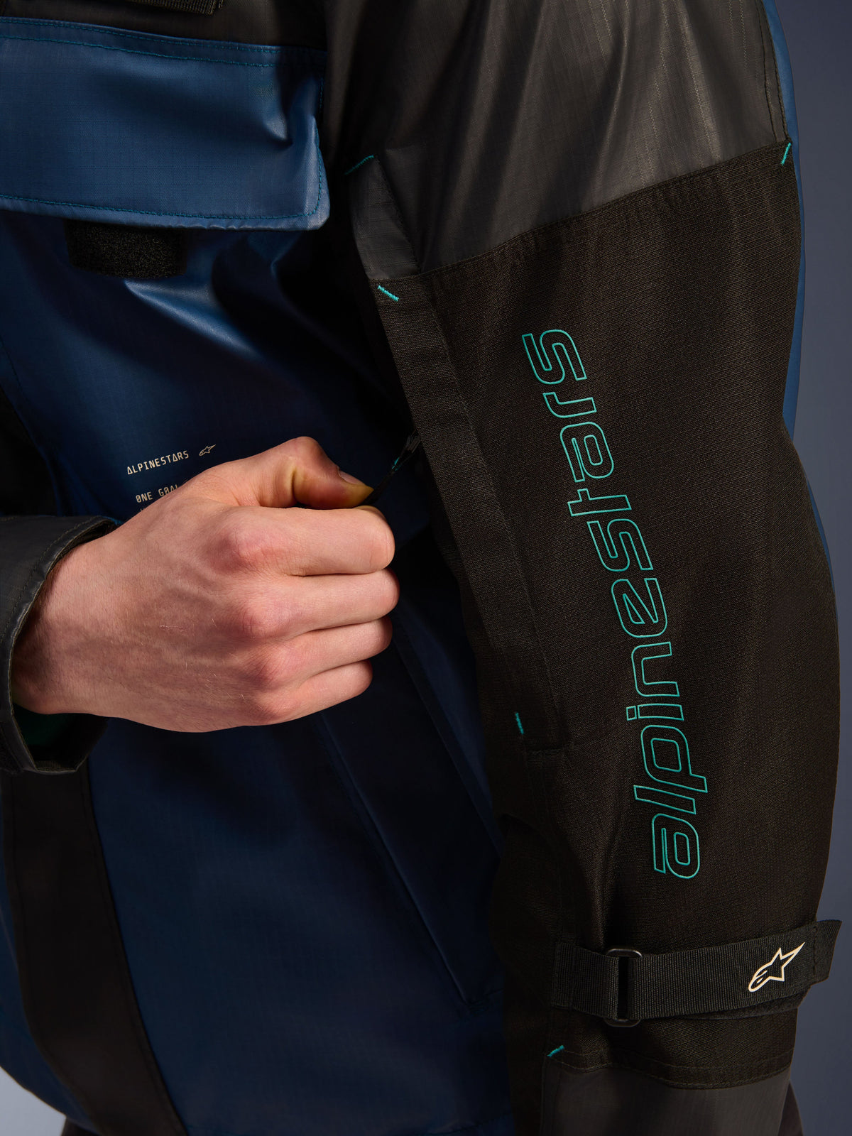 Alpinestars Mospher Waterproof Jas in Black Dark Petrol, close-up detail van de mouw met petrolblauwe Alpinestars-branding, een hand die de ventilatierits aanpast, voorzien van duurzame ripstop-stof en een verstelbare mouwband