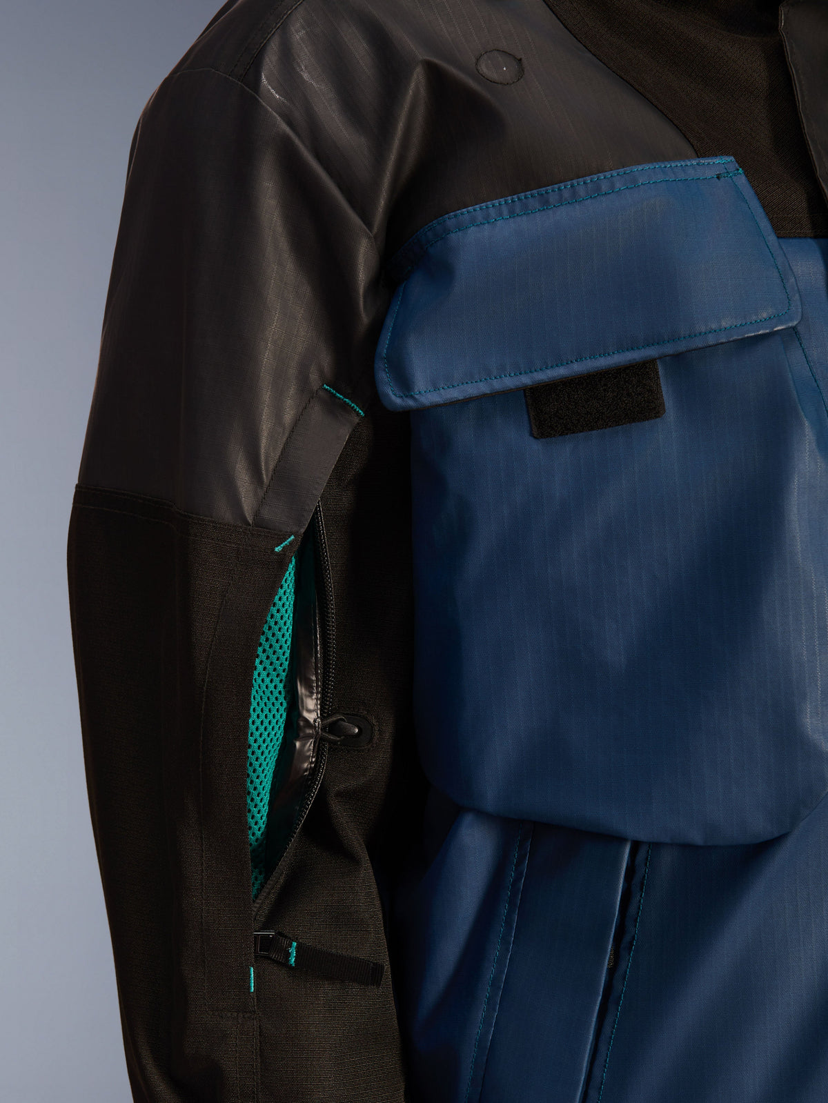 Close-up van Alpinestars Mospher WP-jas in Zwart en Dark Petrol, voorzien van een open ventilatierits op de mouw met felblauwe mesh voering, een grote borstzak en duurzaam ripstop textielmateriaal.