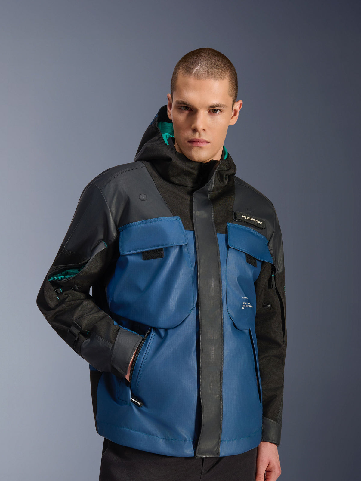Mannelijk model draagt de Alpinestars Mospher Waterproof Jacket in Black en Dark Petrol, met grote utility borstzakken, een ontwerp met capuchon met groenblauwe voering en technische ripstop-stof voor stedelijk woon-werkverkeer op de motor.