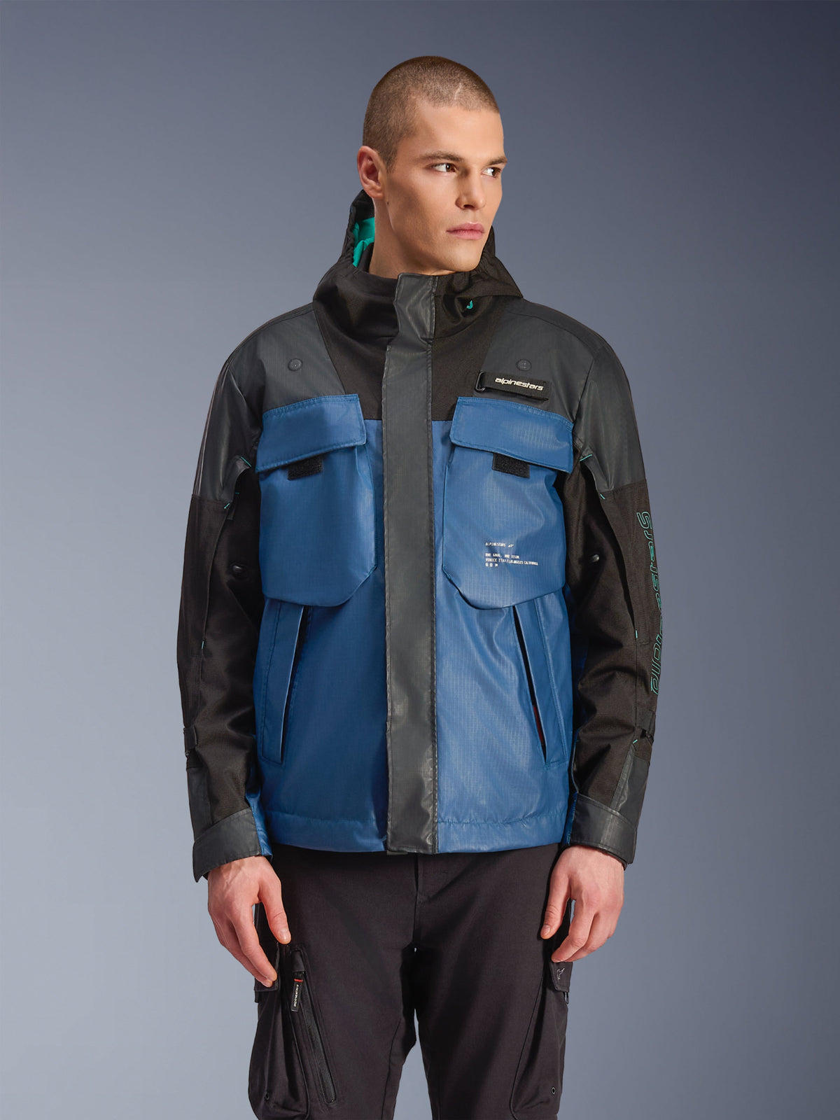 Alpinestars Mospher waterdichte jas, urban motorparka, zwart en donker petrolblauw, met grote tactische borstzakken, hoge kraag met capuchon, technische ripstop-textuur, vooraanzicht op model