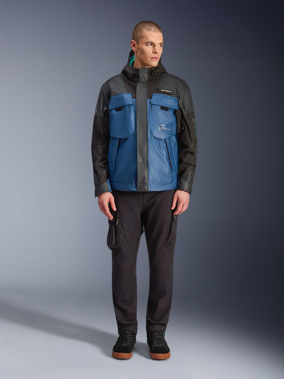 Alpinestars Mospher Waterproof Jacket in Black Dark Petrol, mannelijk model draagt tactisch geïnspireerde all-season motorparka met grote functionele zakken en geïntegreerde capuchon, gecombineerd met een zwarte technische broek en zwarte schoenen, studio vooraanzicht