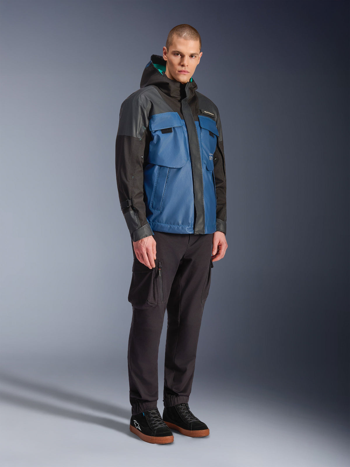 Alpinestars Mospher Waterproof Jacket, zwart en Dark Petrol blauw, gedragen met een zwarte cargobroek en zwarte sneakers, stedelijke forenzenstijl met functionele zakken en capuchon