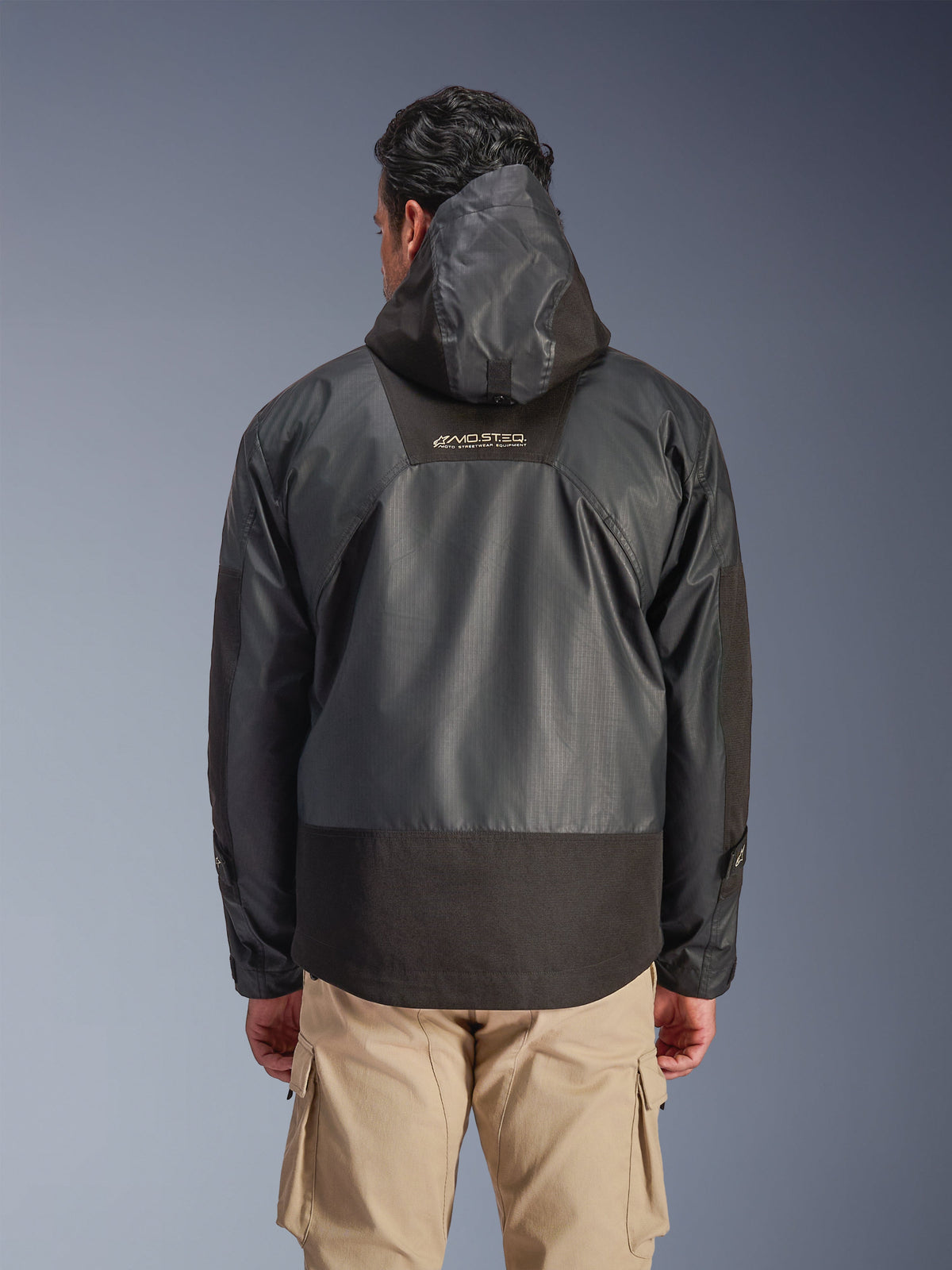 Rückansicht eines Mannes, der die Alpinestars Mospher Waterproof Jacke in Schwarz trägt, mit Kapuze und strapazierfähiger Ripstop-Textilkonstruktion, kombiniert mit beigen Cargohosen, das MO.ST.EQ.-Logo auf dem oberen Rücken hervorhebend, urbanes Motorradbekleidungs-Design.