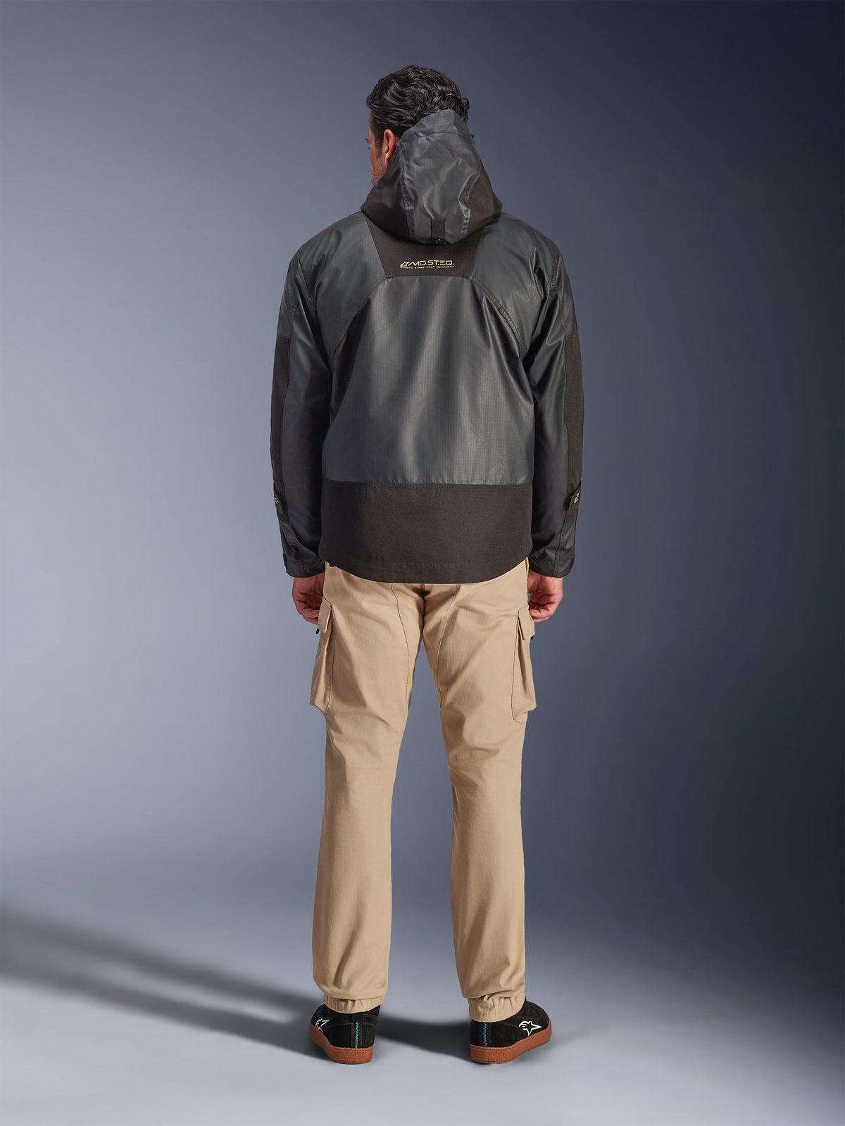 Alpinestars Mospher Waterproof Jacke, schwarzer Motorrad-Parka in der Rückansicht, mit MO.ST.EQ-Logo auf dem oberen Rücken und Ripstop-Gewebe-Konstruktion, getragen mit hellbrauner Cargohose und schwarzen Sneakern, urbaner Pendler-Stil.