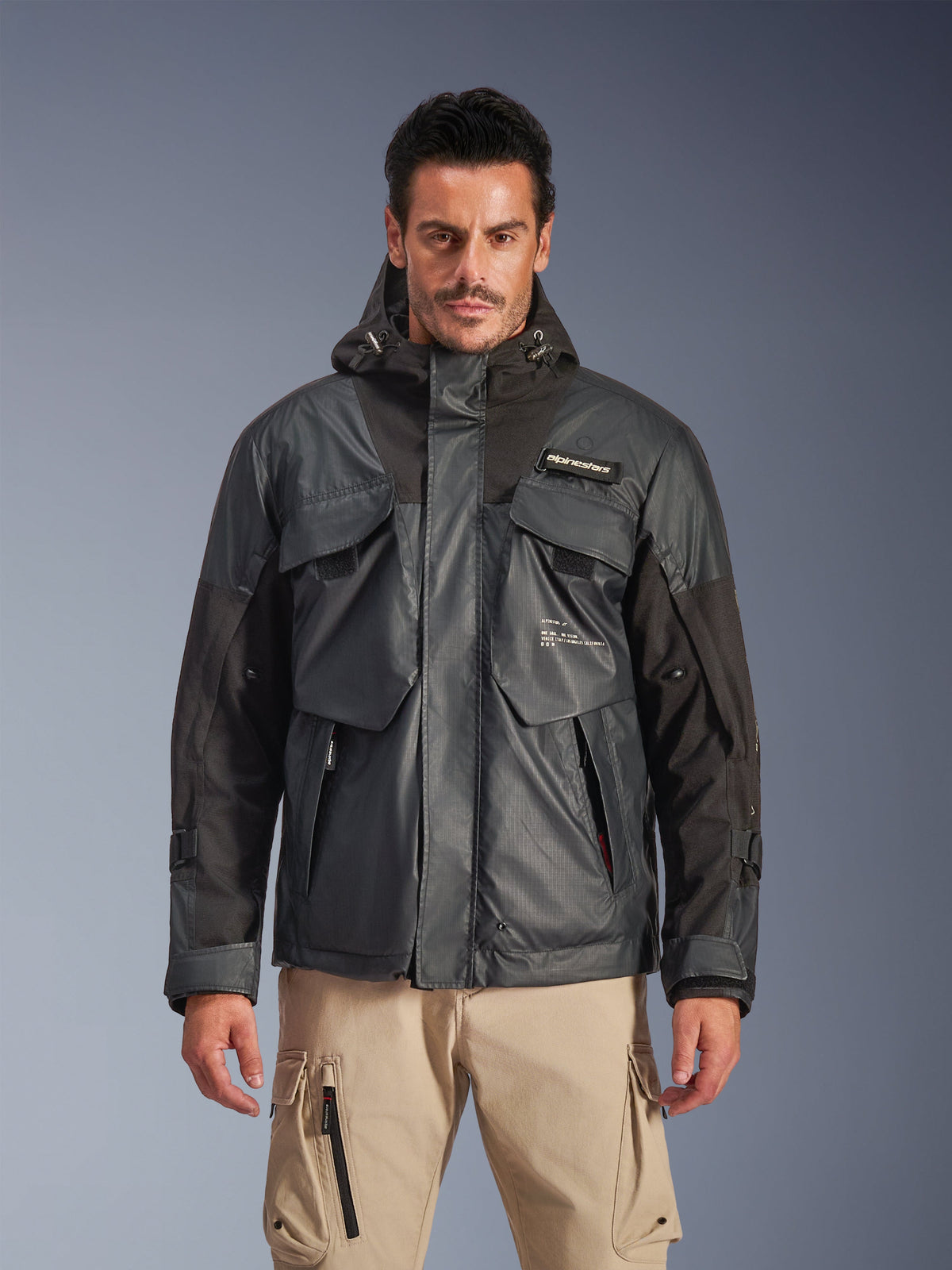 Alpinestars Mospher Waterproof Jacke, Urbaner Motorrad-Parka für Herren, schwarz und dunkelgraues Ripstop-Gewebe, Kapuzendesign mit mehreren großen Funktionstaschen, getragen von einem Modell mit khakifarbenen Cargohosen für einen taktischen Look