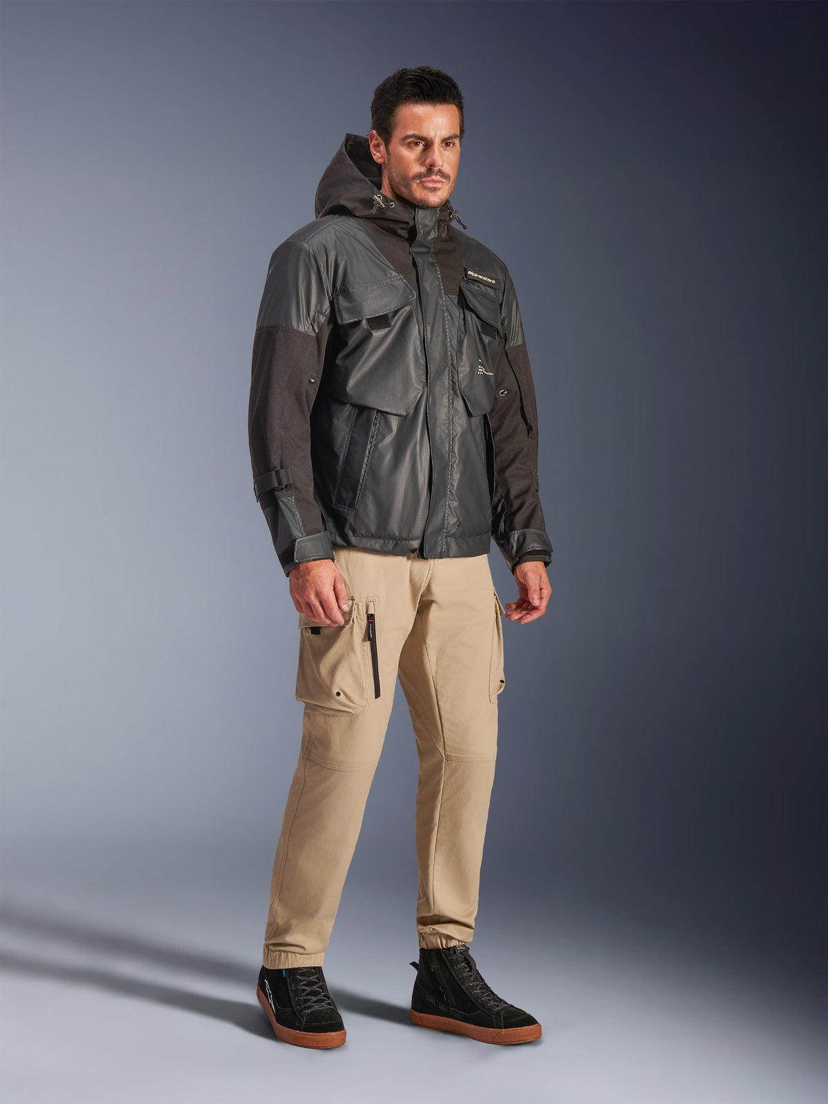 Männliches Modell in der Alpinestars Mospher Waterproof Jacke in Schwarz, wasserdichter Parka im Utility-Look mit Multi-Taschen-Design und Kapuze, kombiniert mit hellbraunen Cargo-Hosen und schwarzen High-Top-Sneakern, urbane Motorrad-Pendlerbekleidung.
