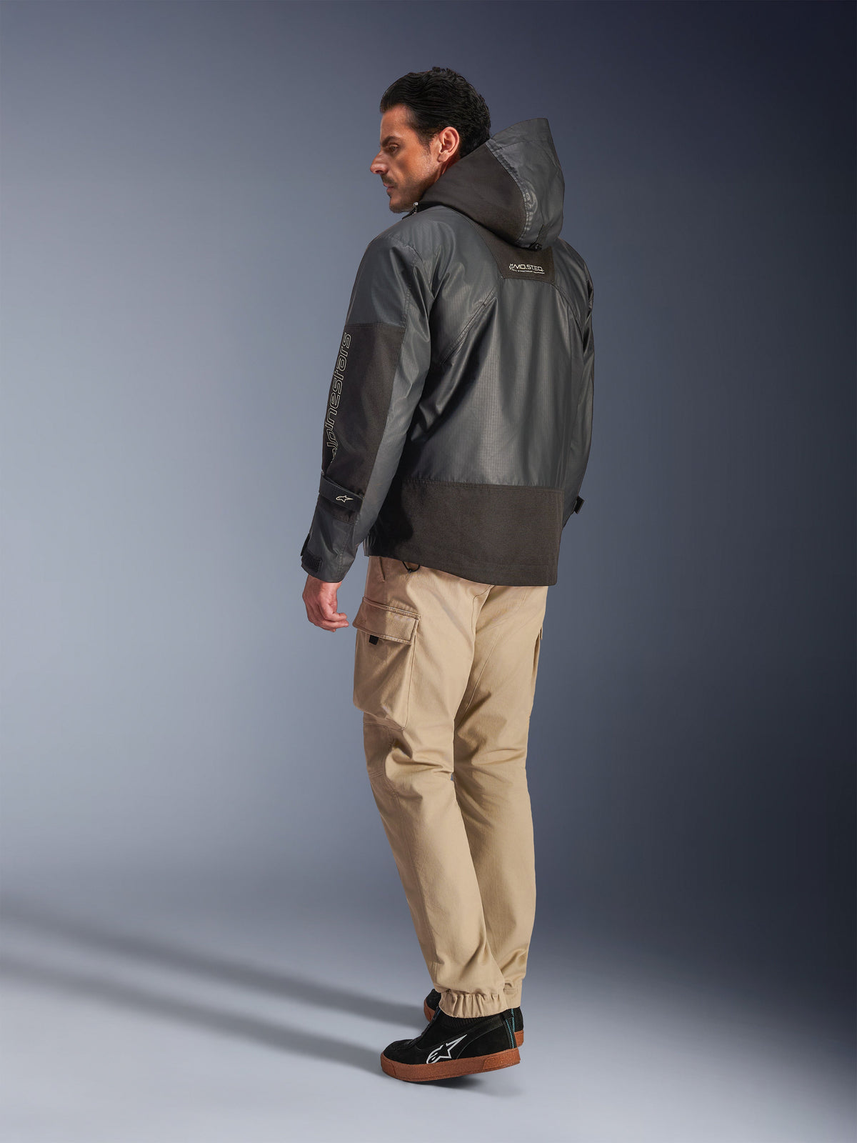 Alpinestars Mospher Waterproof Jacke in Schwarz, Rück- und Seitenprofilansicht, kombiniert mit beigefarbener Cargohose und schwarzen Sneakern, Parka-Design mit Kapuze aus Ripstop-Gewebe, Alpinestars-Branding am Ärmel, moderner urbaner Motorrad-Stil