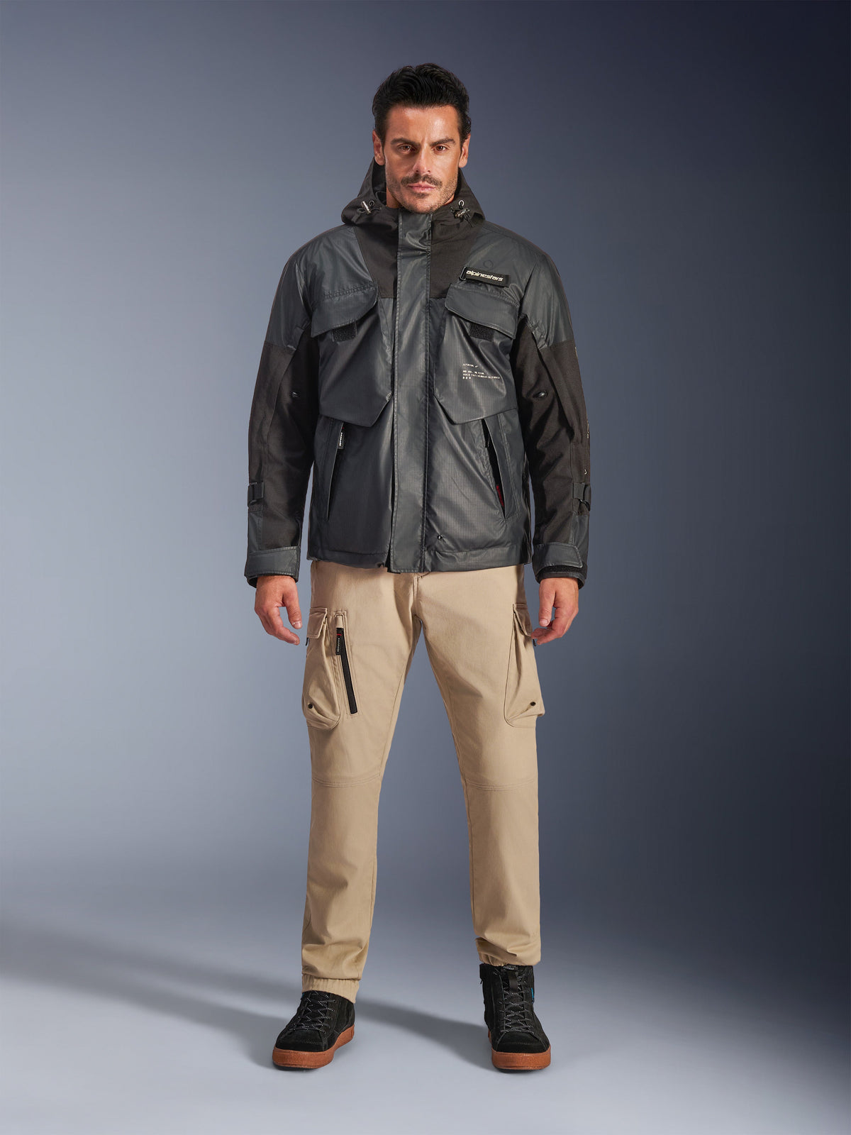 Männliches Model in der Alpinestars Mospher Waterproof Jacke in Schwarz, urbane zeitgemäße Motorradausrüstung, taktisches Utility-Design mit mehreren funktionellen Taschen und Kapuze, kombiniert mit beiger Cargohose und schwarzen Schuhen, Ganzkörper-Studioporträt.