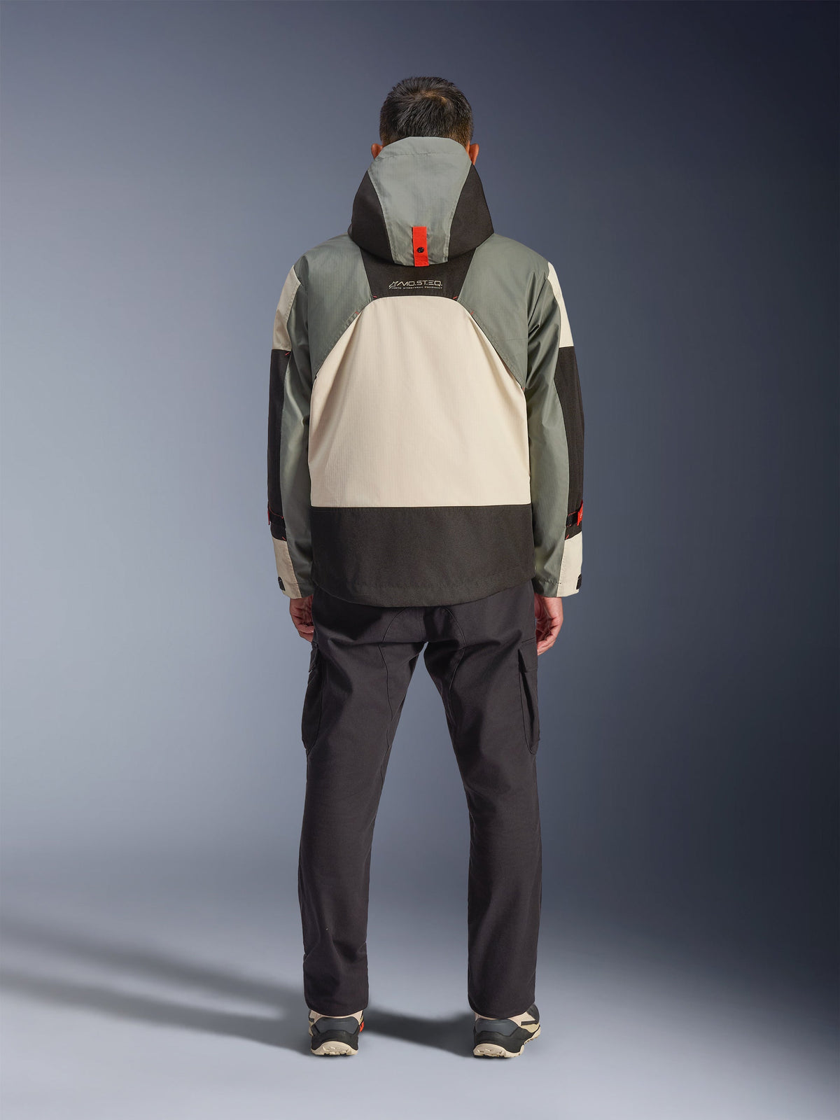 Vue de dos d'un motard portant la veste imperméable Alpinestars Mospher en Fur Gray Black Ice, parka d'inspiration tactique avec design color-block gris, noir et crème, dotée d'une capuche avec sangle orange, associée à un pantalon cargo noir.