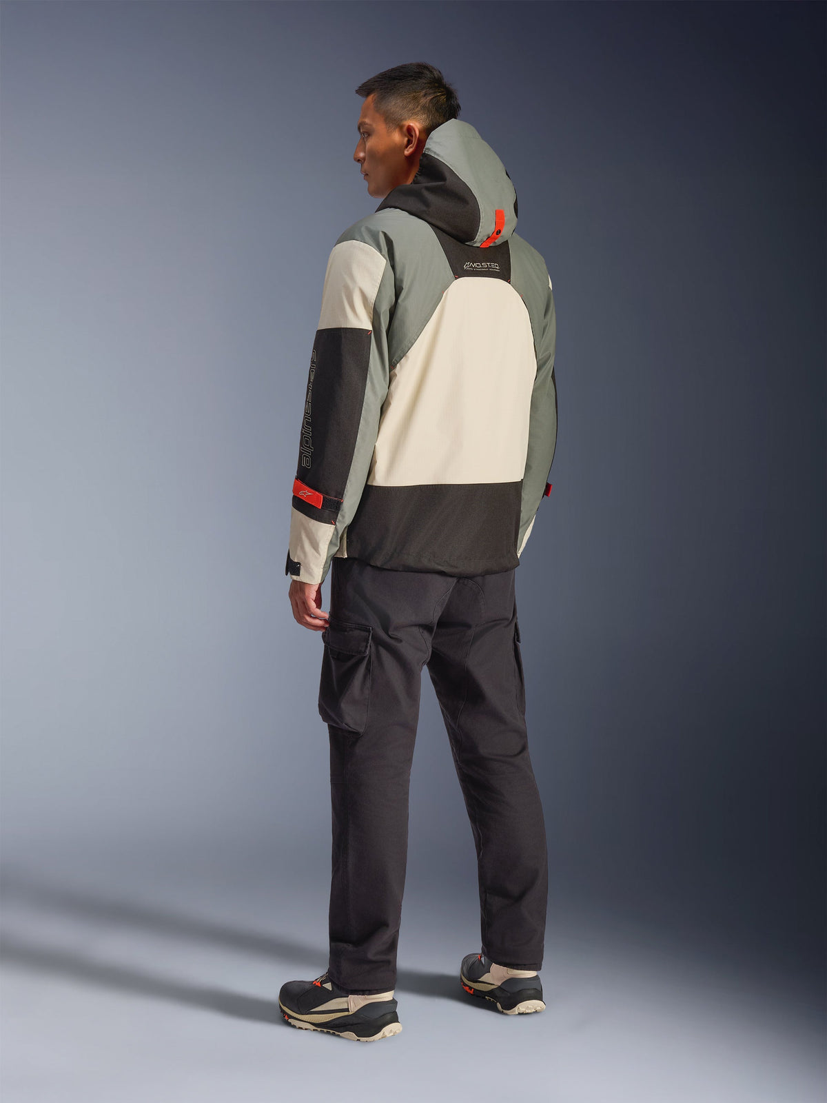 Veste imperméable Alpinestars Mospher en Fur Gray Black Ice, vue arrière d'un mannequin portant une parka de moto tactique avec des empiècements crème, gris et noirs, dotée d'accents rouges et d'une capuche, associée à un pantalon cargo noir et des bottes techniques