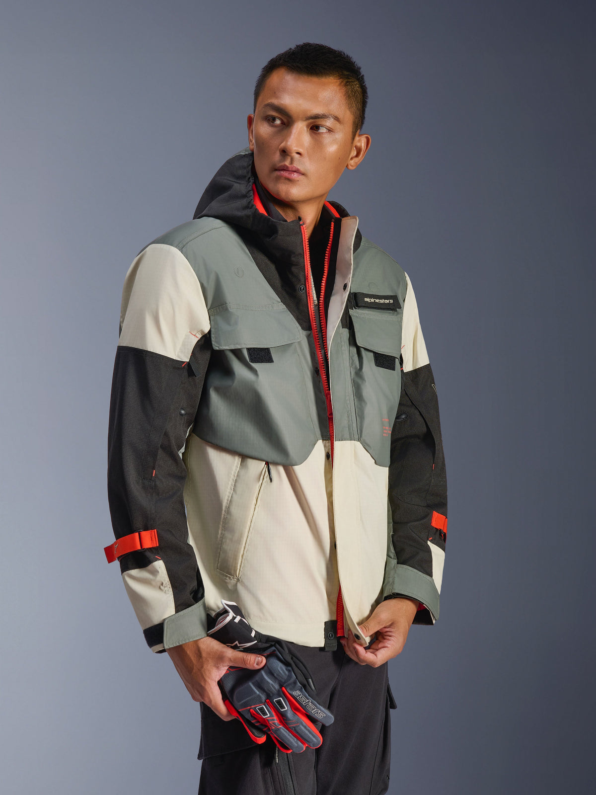 Veste imperméable Alpinestars Mospher en Fur Gray Black Ice, parka textile multi-panneaux avec sections grises, noires et blanc cassé, accents de fermetures éclair et sangles rouge vif, design d'inspiration tactique avec capuche et poches utilitaires, portée par un mannequin tenant des gants de moto.