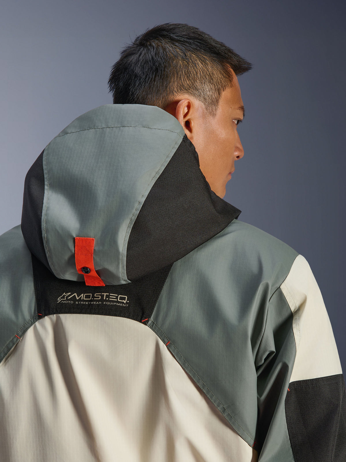 Veste imperméable Alpinestars Mospher, Fur Gray Black Ice, vue arrière rapprochée montrant le système d'ancrage de la capuche avec une languette à bouton-pression orange, marquage MO.ST.EQ. Moto Streetwear Equipment, construction en textile ripstop durable