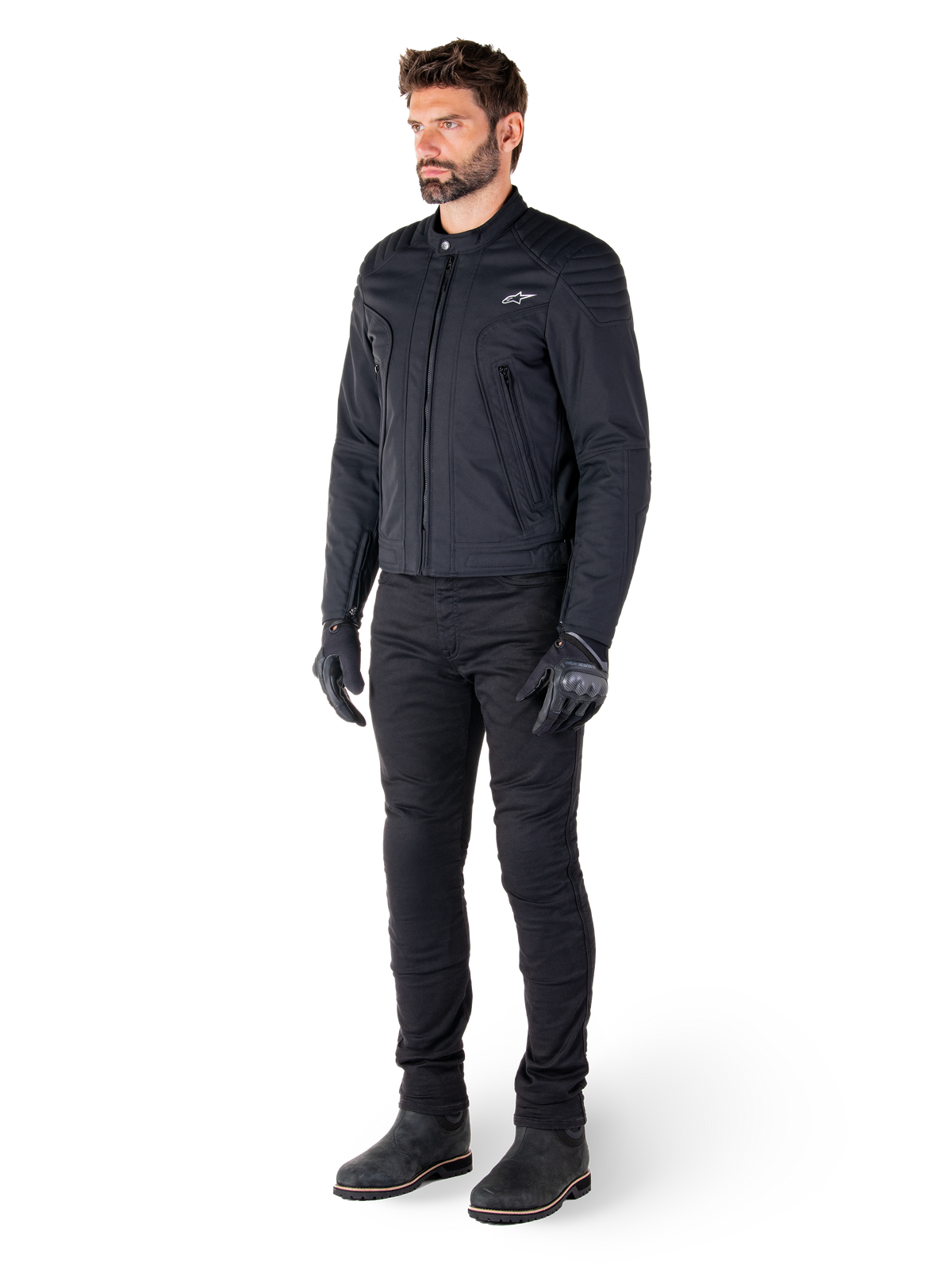 Pilote portant la veste Clayton WR d'Alpinestars, veste de moto urbaine noire, coupe ajustée avec détails matelassés aux épaules et finition mate, associée à un pantalon technique et des gants de moto noirs.