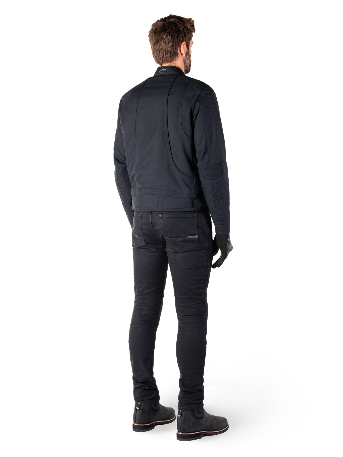 Vue arrière d'un motard portant la veste Alpinestars Clayton WR en noir, avec empiècements matelassés aux coudes et aux épaules, coutures ergonomiques incurvées et détails réfléchissants au col, associée à un pantalon et des bottes de moto noirs pour un look urbain.