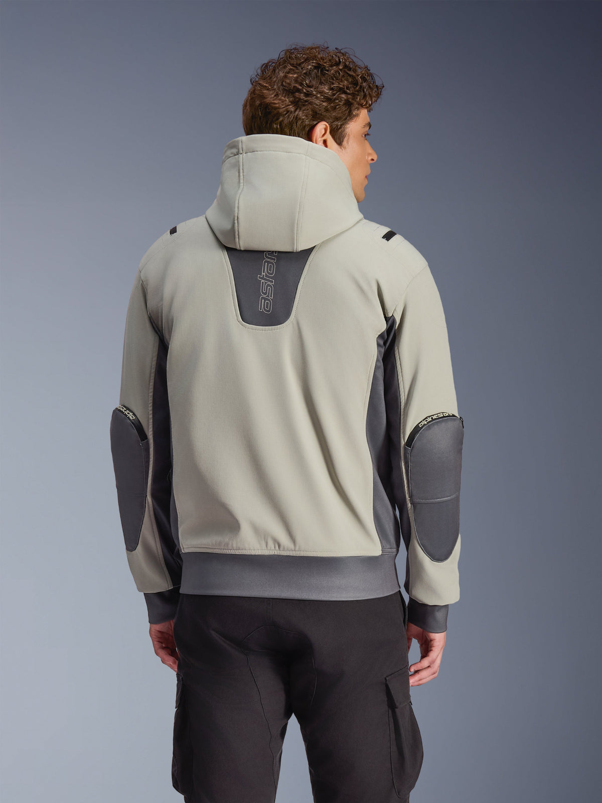 Alpinestars Moblast Wasserdichte Jacke in Iron Gray und Dark Sage, Rückansicht am Model, urbane Motorradjacke mit Kapuze, dunkelgraue Stretcheinsätze und verstärkte Ellbogen, kombiniert mit schwarzer Cargohose