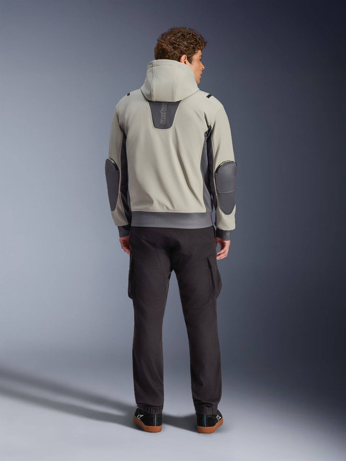 Alpinestars Moblast Wasserdichte Jacke, Iron Gray Dark Sage, Rückansicht eines männlichen Fahrers in der Motorradjacke mit Kapuze, Farbkombination aus Hellgrau und Dark Sage, vertikales „astars“-Logo am oberen Rücken, dunkelgraue Ellbogen-Patches, kombiniert mit dunkler Cargohose für einen modernen urbanen Look.