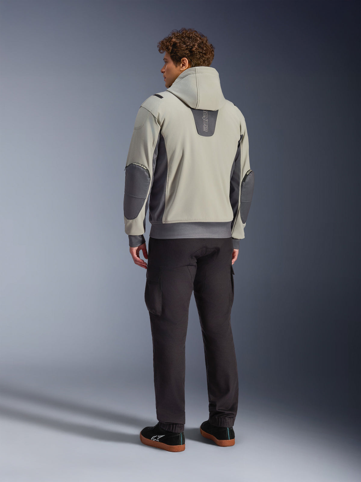 Männliches Model in der Rückansicht, trägt die Alpinestars Moblast Waterproof Jacke in Iron Gray Dark Sage, urbane Motorradjacke mit Kapuze, dunkelgrauen Seiteneinsätzen und verstärkten Ellbogen-Patches, kombiniert mit dunkler Motorradhose und schwarzen Sneakern, moderne minimalistische Ästhetik für den urbanen Pendelverkehr.
