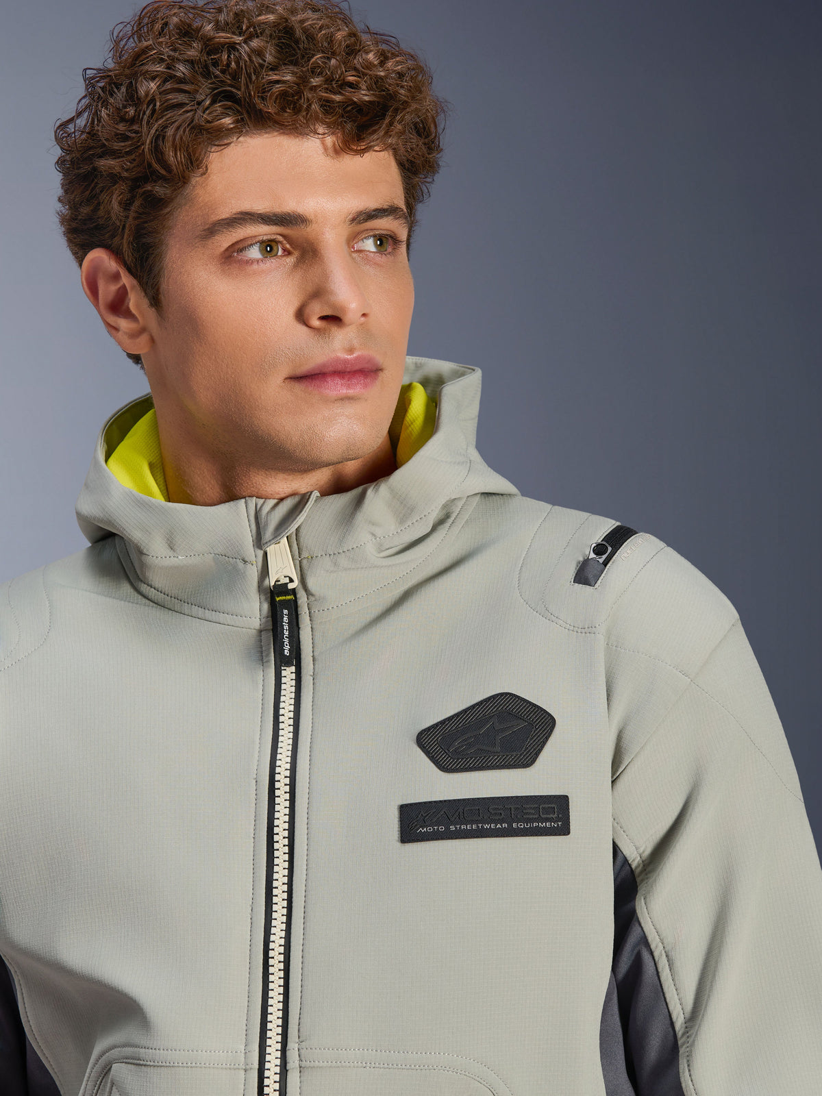 Alpinestars Moblast Wasserdichte Jacke, Iron Gray Dark Sage, Nahaufnahme eines männlichen Models, das die Jacke trägt, mit schwarzem Alpinestars-Logo und MO.ST.EQ.-Brustaufnähern, leuchtend gelbem Kapuzenfutter und einem modernen technischen Design.