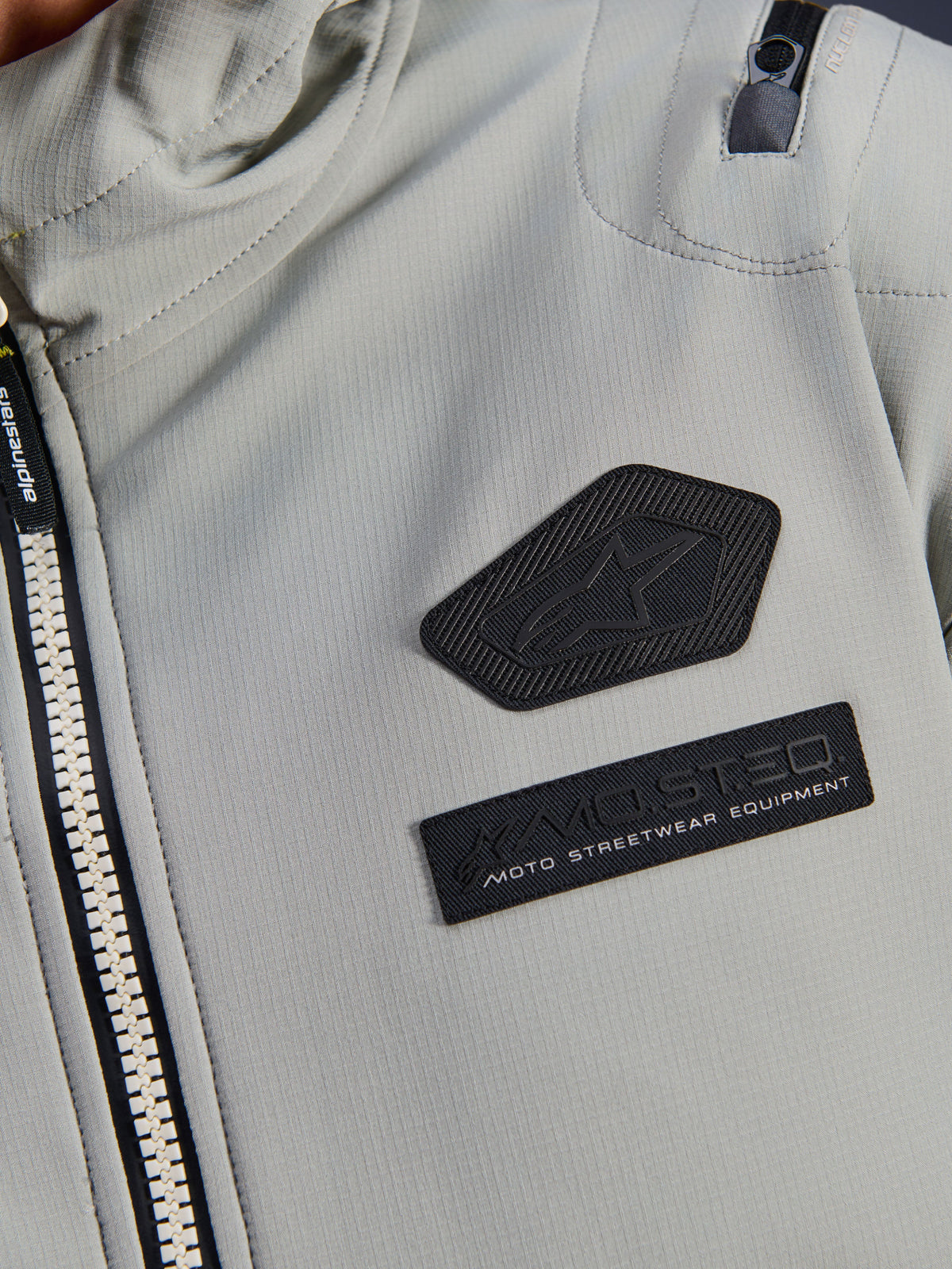 Alpinestars Moblast Wasserdichte Jacke, Iron Gray, Nahaufnahme der Brustaufnäher mit dem Alpinestars Logo und MO.ST.EQ. Moto Streetwear Equipment Branding, technisches Ripstop-Gewebe mit robustem Frontreißverschluss und Detail der Schultertasche.