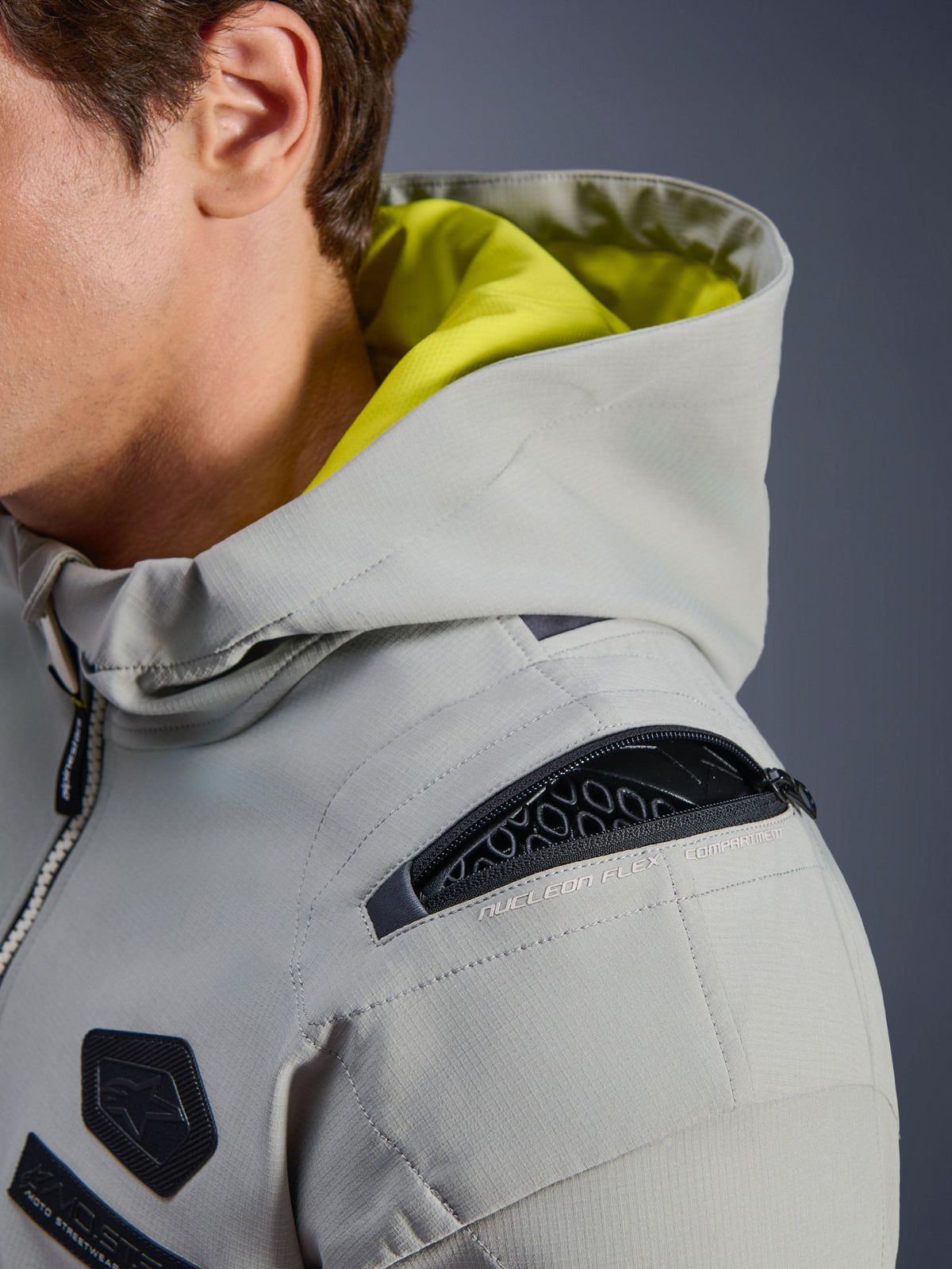 Alpinestars Moblast WP Jacke, Iron Gray, Nahaufnahme der Schulter mit offenem Nucleon Flex Protektorenfach, neongelbes Kapuzeninnenfutter, technische urbane Motorradausrüstung mit funktionellen Reißverschlüssen
