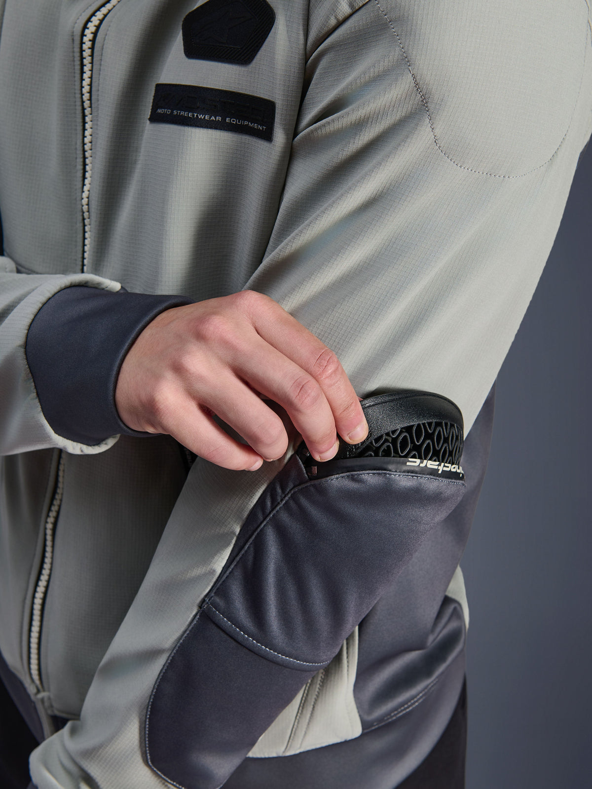Alpinestars Moblast Wasserdichte Jacke, Iron Gray und Dark Sage, Nahaufnahme der Entnahme des Nucleon Flex Plus Protektors aus der äußeren Ellenbogen-Reißverschlusstasche, technische Textil-Urban-Motorradausrüstung mit MO.ST.EQ. Branding