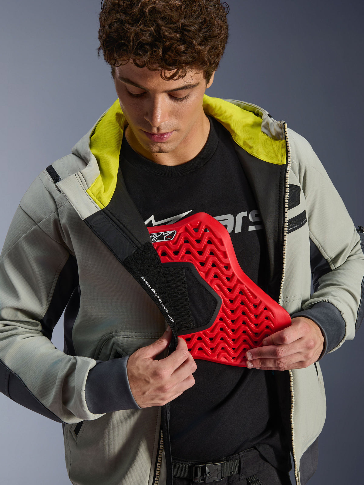 Alpinestars Moblast wasserdichte Jacke, Iron Gray und Dark Sage, Modell demonstriert die Integration des Brustschutzes mit einem roten Protektor-Einsatz, helles limettengrünes Innenfutter, moderne urbane Motorradbekleidung