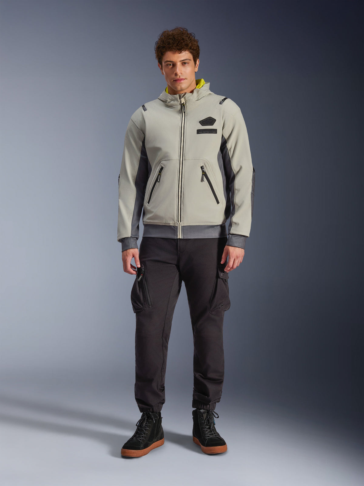 Männliches Model in Alpinestars Moblast Waterproof Jacke in Iron Gray und Dark Sage, Motorradjacke mit Kapuze und modernem technischen Design, kombiniert mit dunkler Cargo-Fahrhose und schwarzen Stiefeln, urbane zeitgenössische Motorsportbekleidung