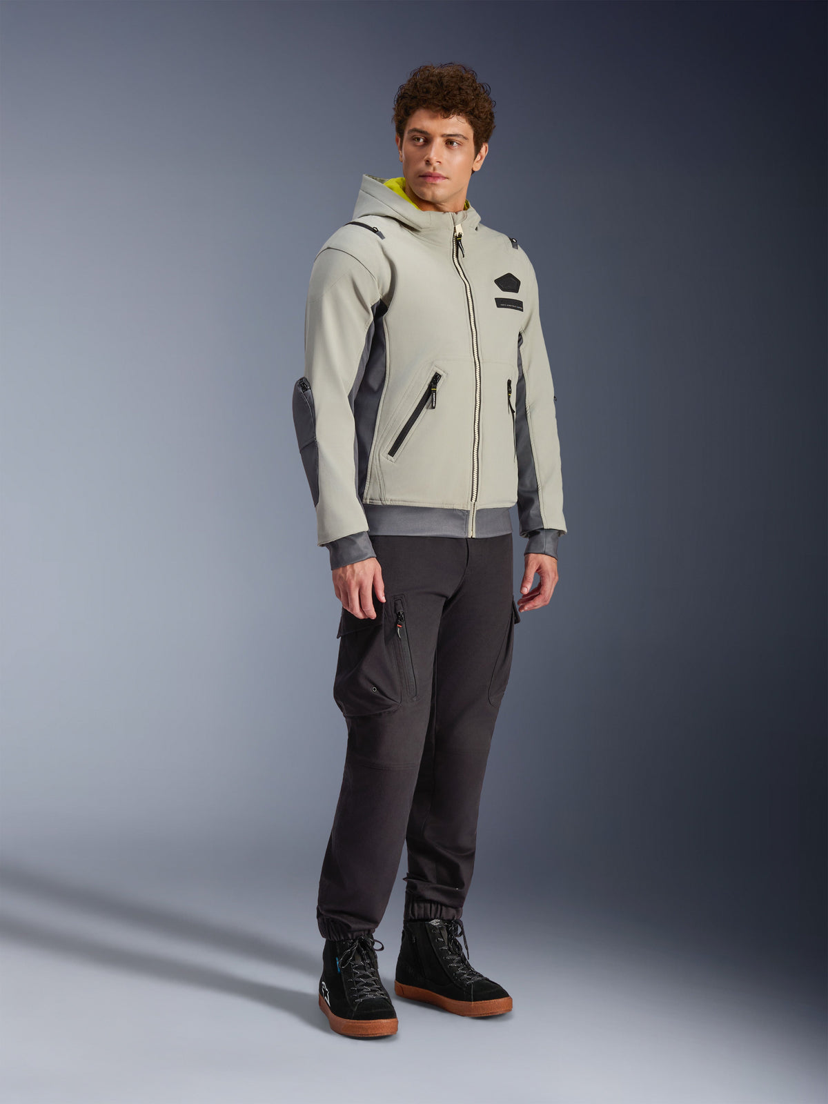 Alpinestars Moblast Wasserdichte Jacke, Iron Gray und Dark Sage, urbane Motorradjacke im Hoodie-Design, getragen von einem Model mit dunkler Motorradhose und schwarzen Stiefeln, minimalistische Cyber-Tech-Ästhetik mit gelber Kapuzeninnenseite und wasserdichten Reißverschlüssen