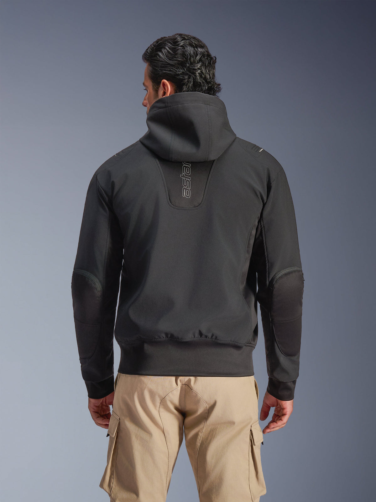 Alpinestars Moblast Wasserdichte Jacke, Schwarz, Rückansicht einer urbanen Motorradjacke mit Kapuze, mit vertikalem 'astars'-Logo auf dem Rücken, elastischem Bund und Bündchen, verstärkten Ellbogen und modernem, technikorientiertem Design, getragen mit einer hellbraunen Cargo-Hose.