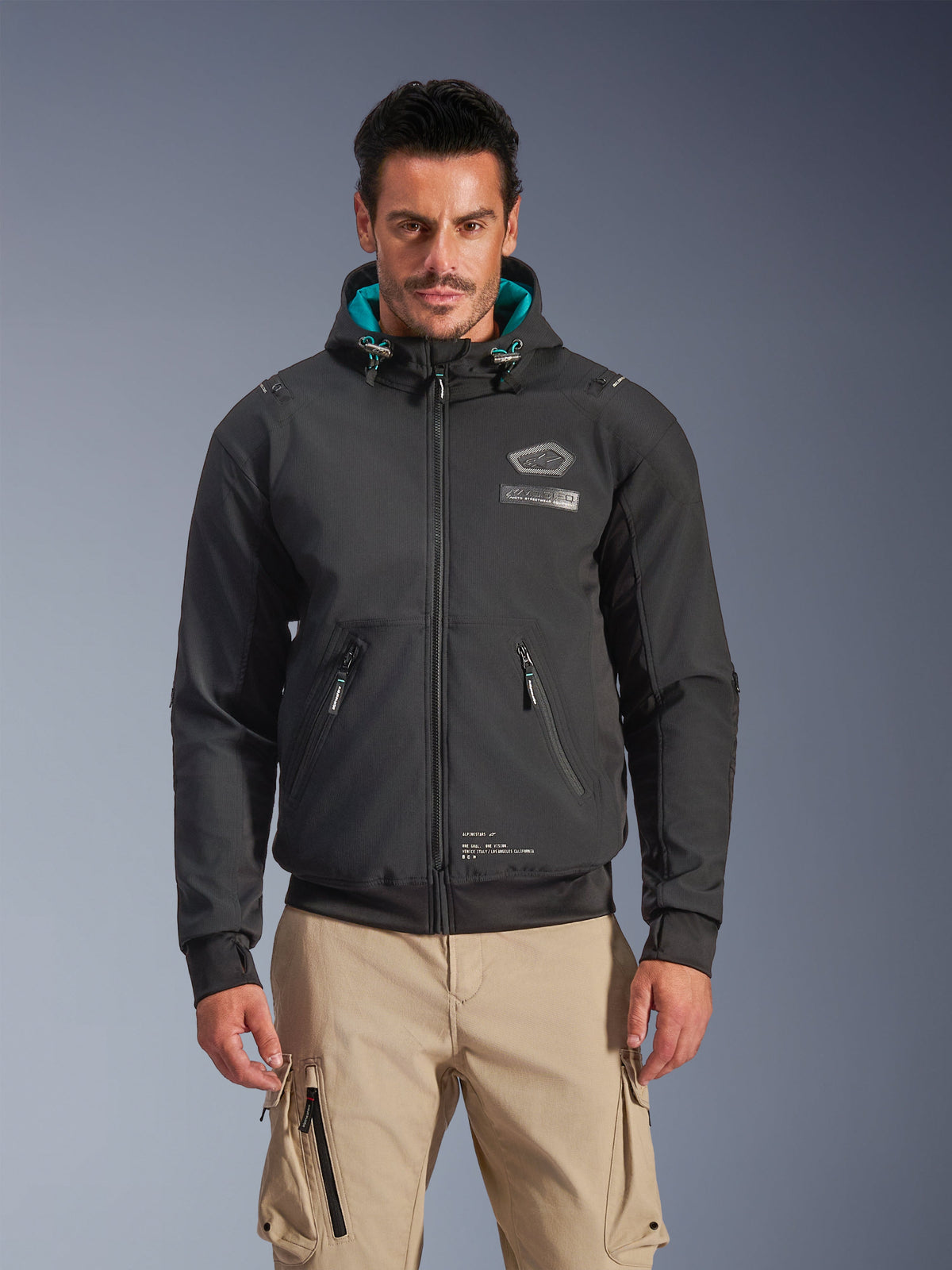 Alpinestars Moblast Wasserdichte Jacke, Urban-Motorradjacke für Herren, Schwarz mit türkisfarbenem Kapuzenfutter, mit 3D-Reißverschlusstaschen und Kapuzendesign, getragen mit beigen Cargohosen, moderne urbane Motorradbekleidung