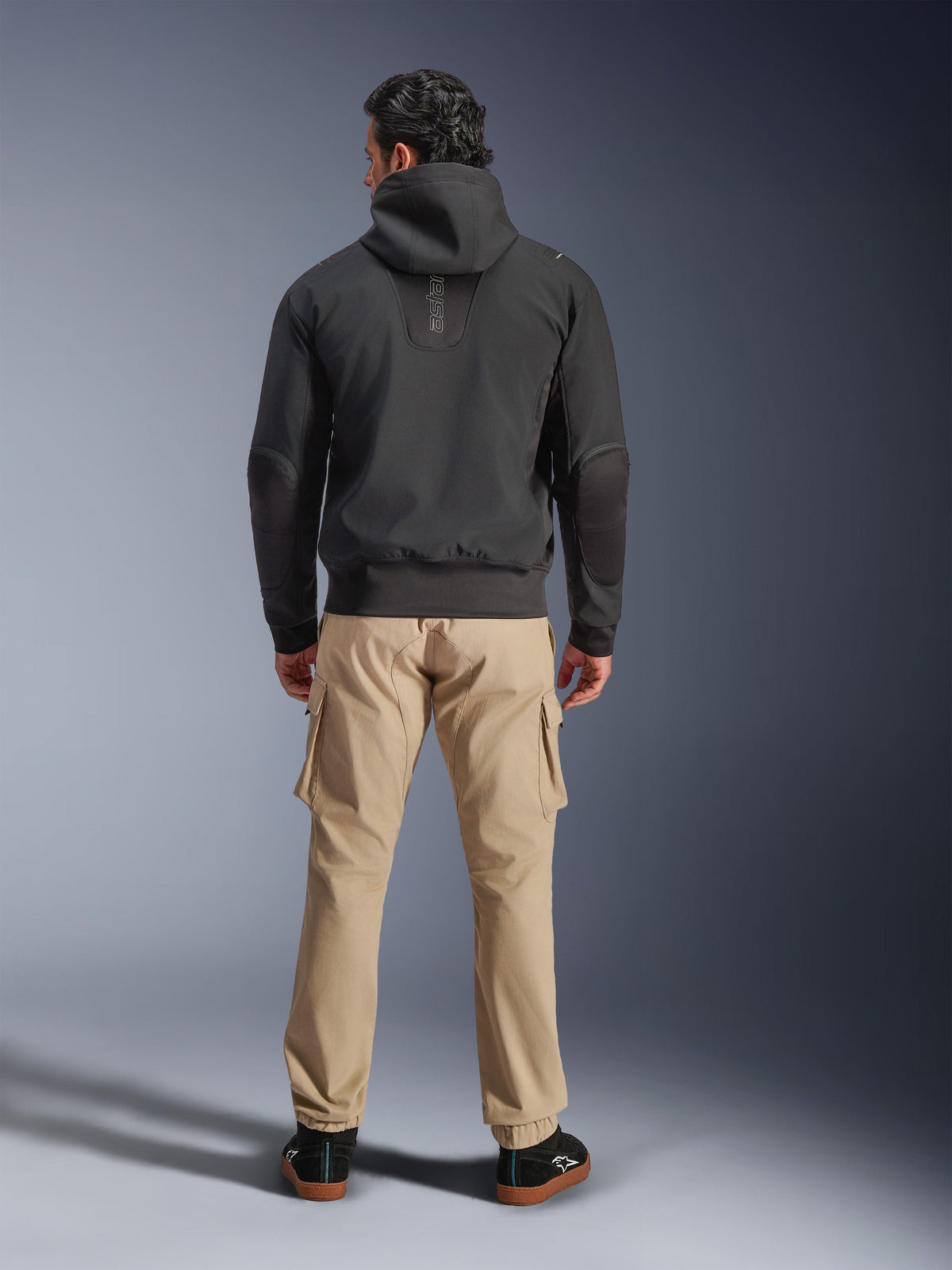 Alpinestars Moblast Wasserdichte Jacke, Schwarz, Rückansicht eines urbanen Motorrad-Hoodies, getragen mit beigen Cargohosen und schwarzen Sneakern, minimalistisches Design mit vertikalem „astars“-Branding am oberen Rücken, moderne Allwetter-Fahrausrüstung